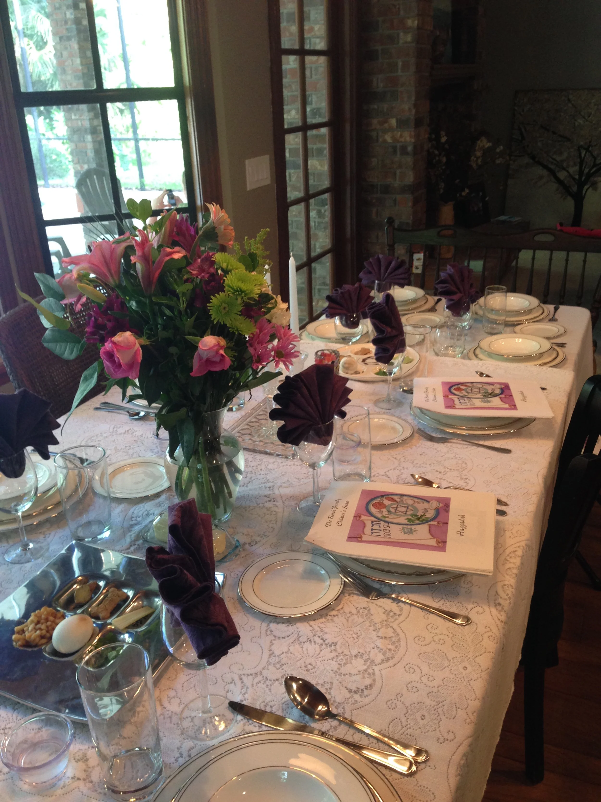 A beautifully set dining table with a lace tablecloth, floral centerpiece, multiple plates, glasses, purple napkins, and printed menus, near windows and a brick wall in a cozy dining room.