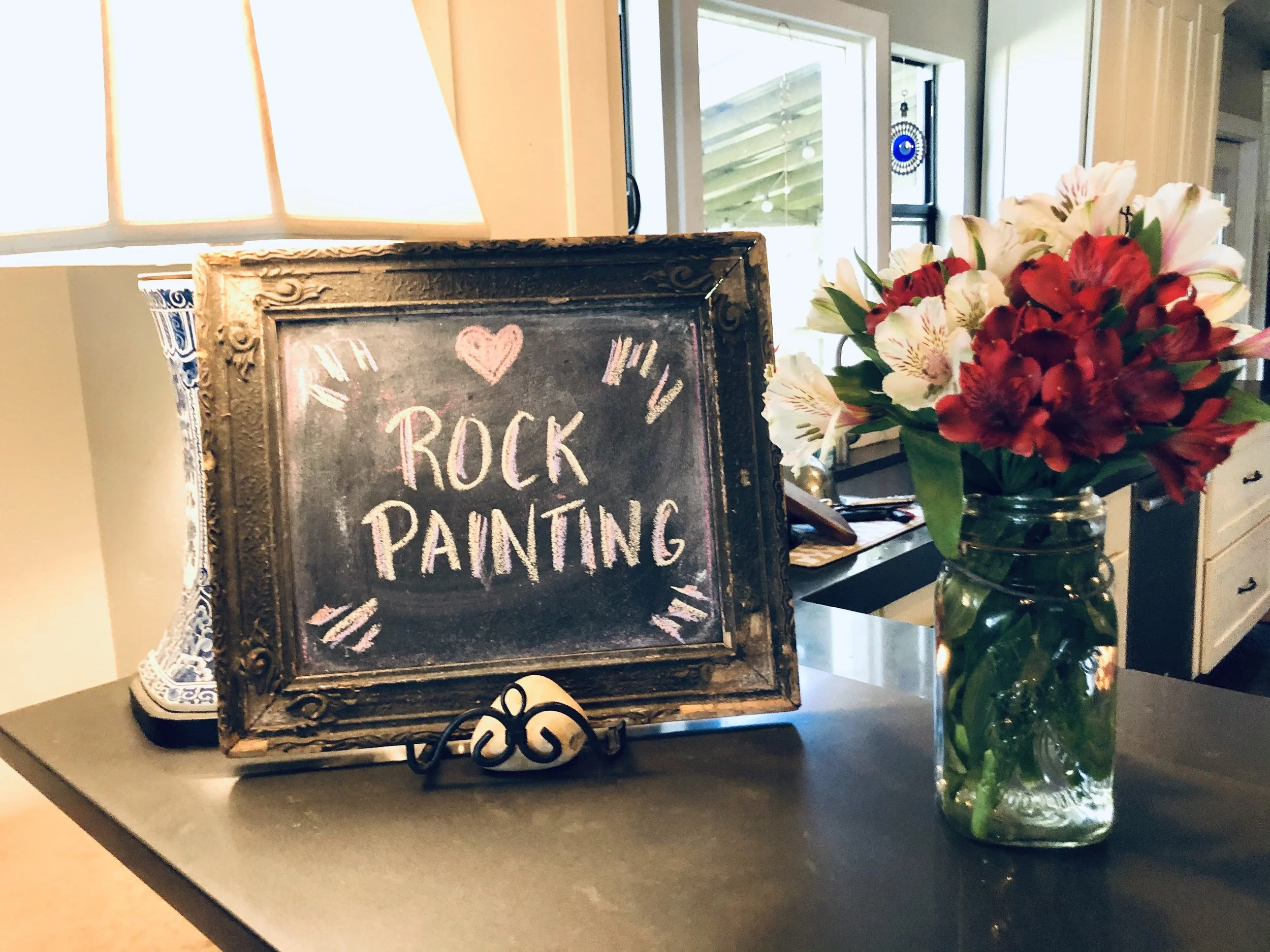 A framed sign on a tabletop reads 'Rock Painting' with a pink heart and decorative accents. Next to the sign is a glass jar filled with pink, white, and red flowers.
