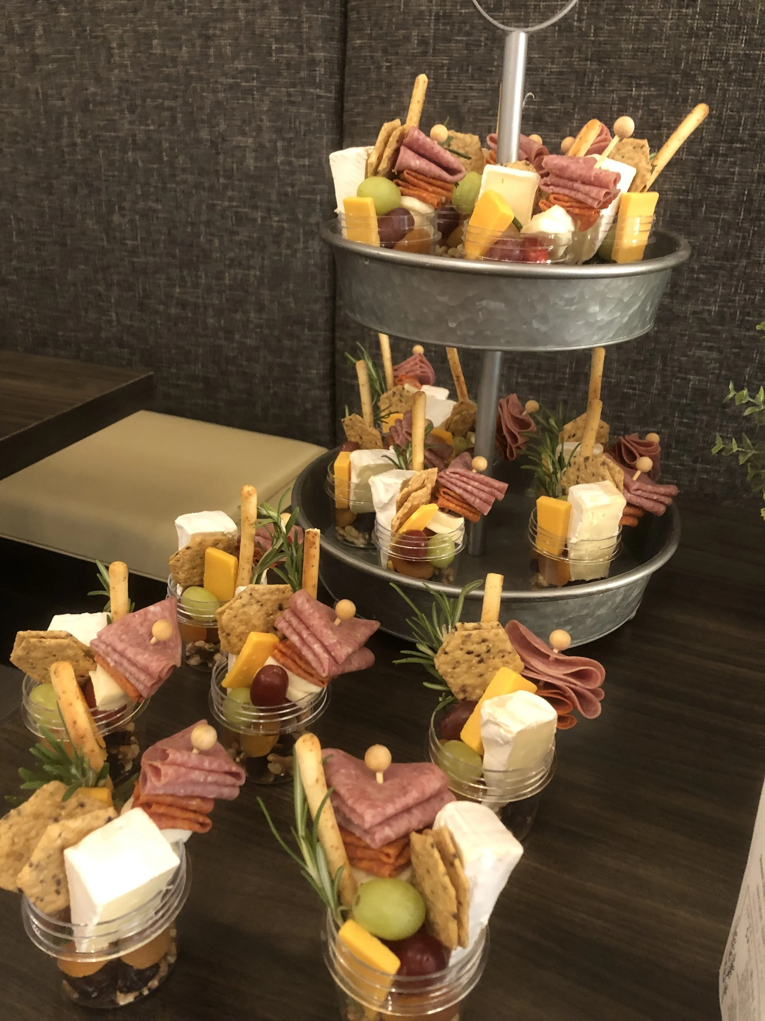 Assorted individual snack cups containing cheese, meats, crackers, grapes, and garnishes, arranged on a dark wooden table in front of a multi-tiered metal stand.