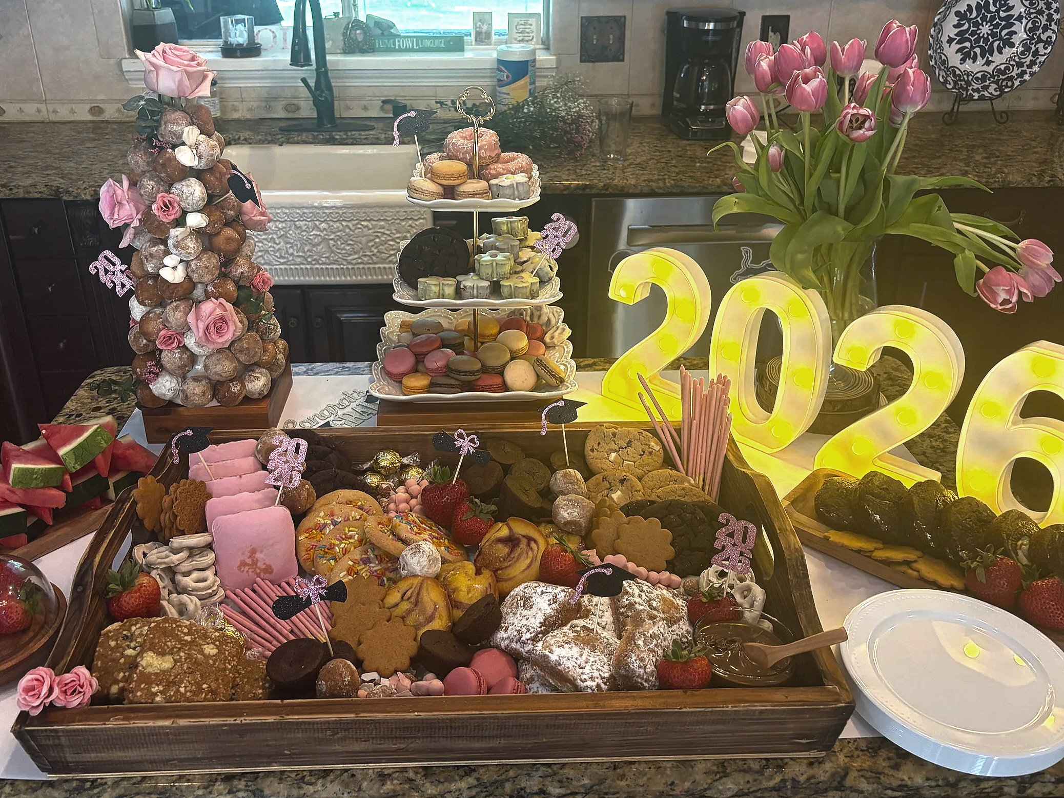 Table with a variety of desserts including macarons, cookies, strawberries, and pastries decorated for a celebration, with pink tulips in a vase and illuminated number '2023' decorations.