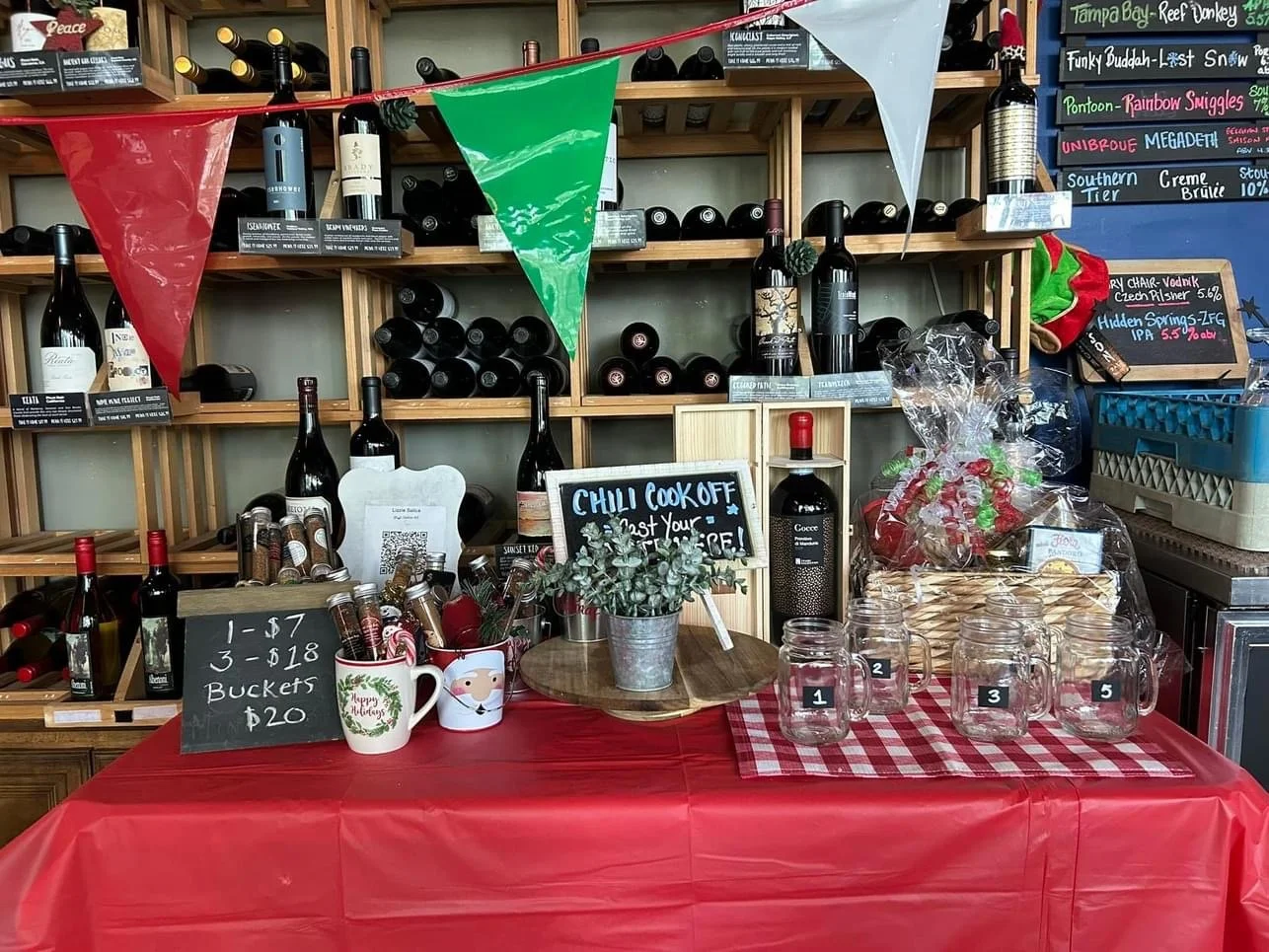 A wine display table decorated for Christmas with a red tablecloth, Mason jars labeled 1, 2, 3, and 5, festive mugs, and a chalkboard sign promoting a chilli cook-off with the message "Eat Your Heart Out!". Banners and gift baskets are also part of t
