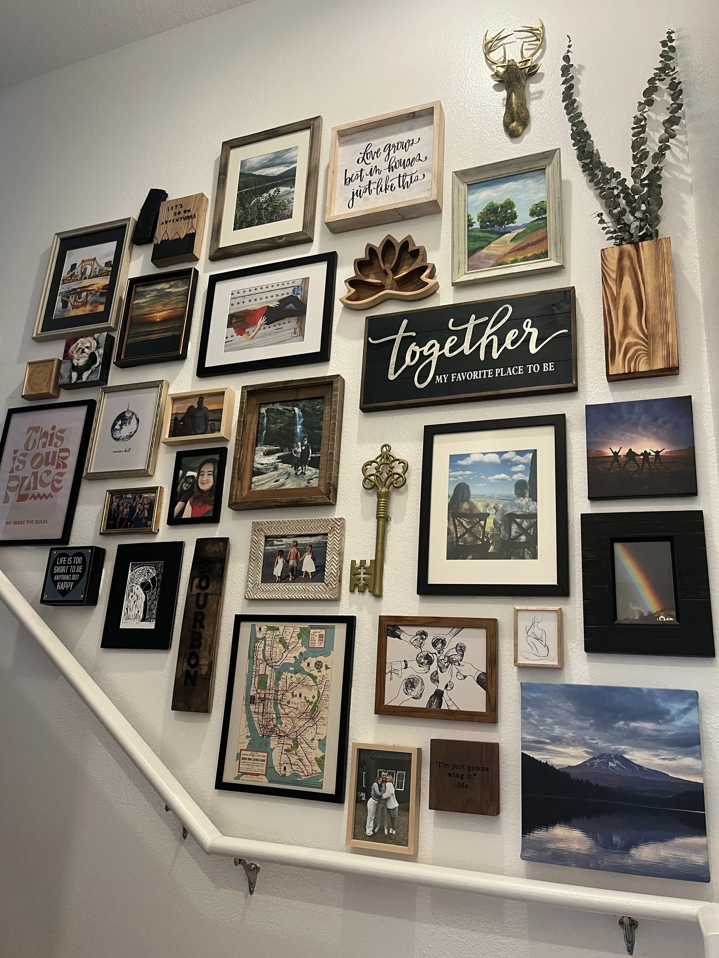 A gallery wall with various framed photos, artwork, and decorative signs arranged Amid a stair railing, including landscape photos, portraits, a rainbow, a map, a key, and text-based art.
