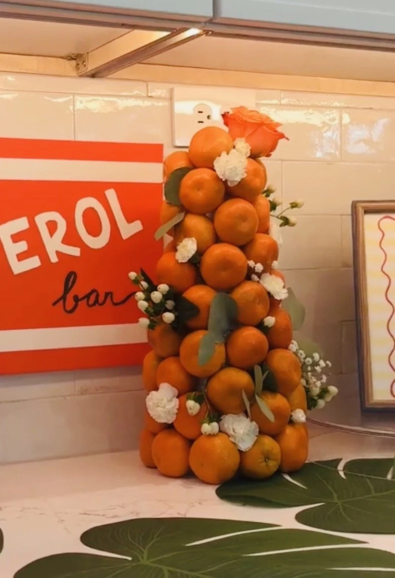 A decorative arrangement of oranges, green leaves, and white flowers, shaped like a small Christmas tree on a counter, with a sign reading 'EROL bar' and a framed sign in the background.