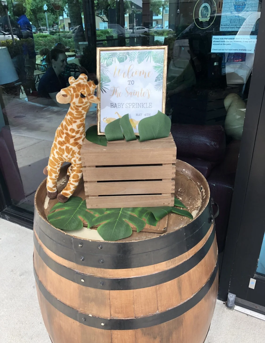 A stuffed giraffe toy standing next to a wooden crate with green leaves around a sign that reads 'Welcome to The Saints Baby Sprinkle May 4th.' The display is arranged on top of a large wooden barrel outside a restaurant or cafe.