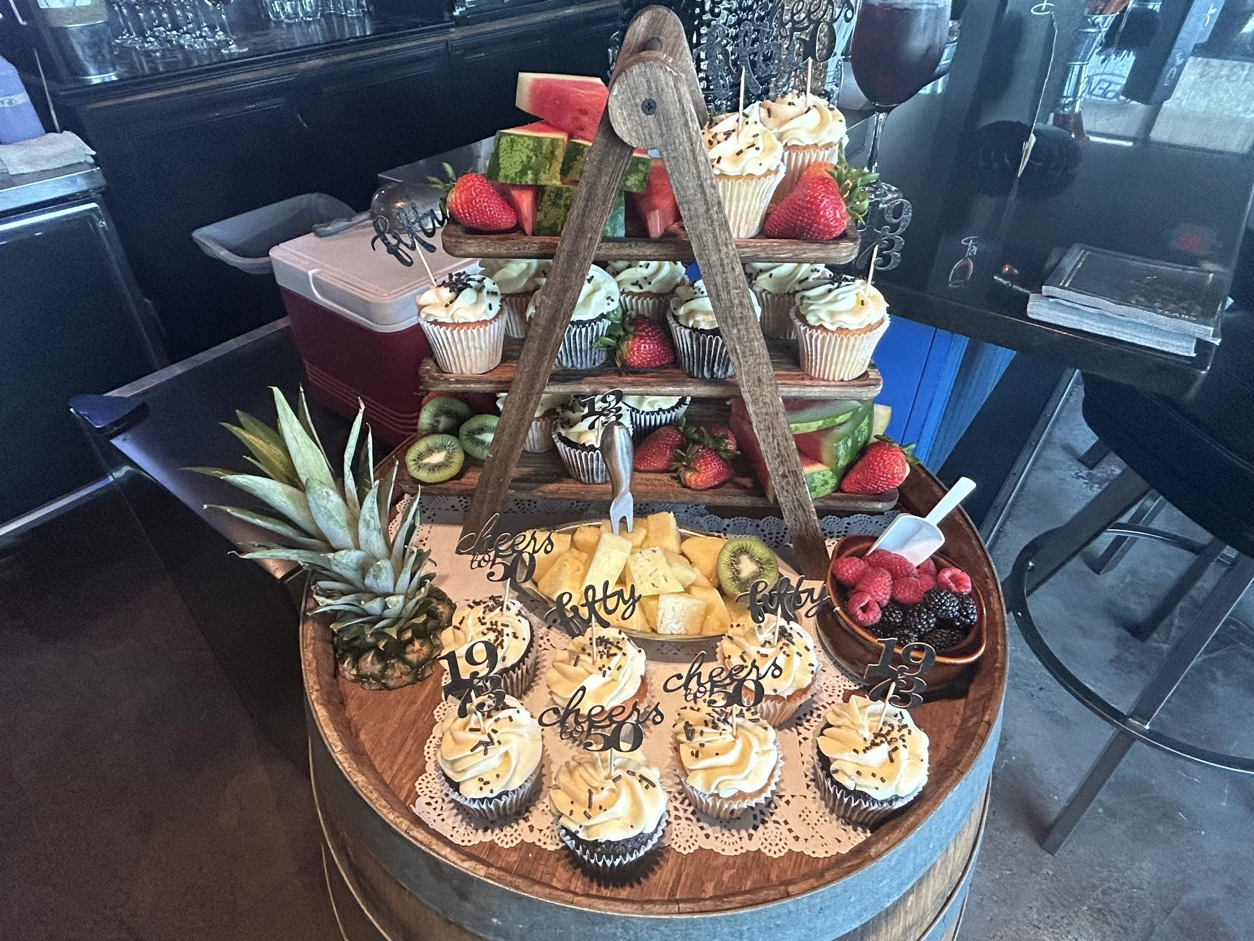 A wooden barrel table with a pineapple, a bowl of berries, a cheese platter, and various cupcakes and fruit arranged on a three-tiered wooden stand, with flags indicating 'cheers' and 'happy birthday'