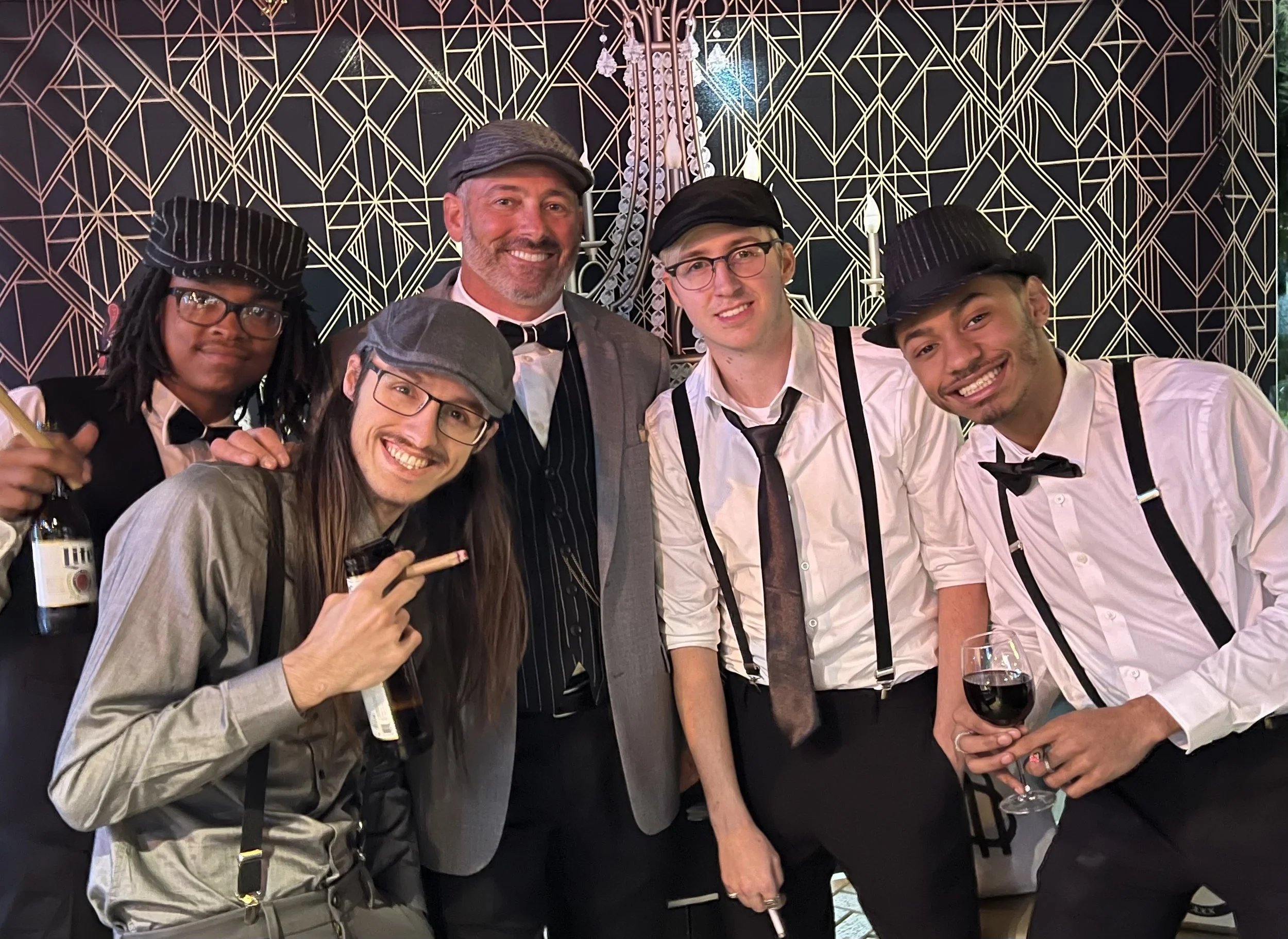 A group of six men in vintage attire, smiling and posing together at a themed event, with a decorative black and gold geometric patterned background.