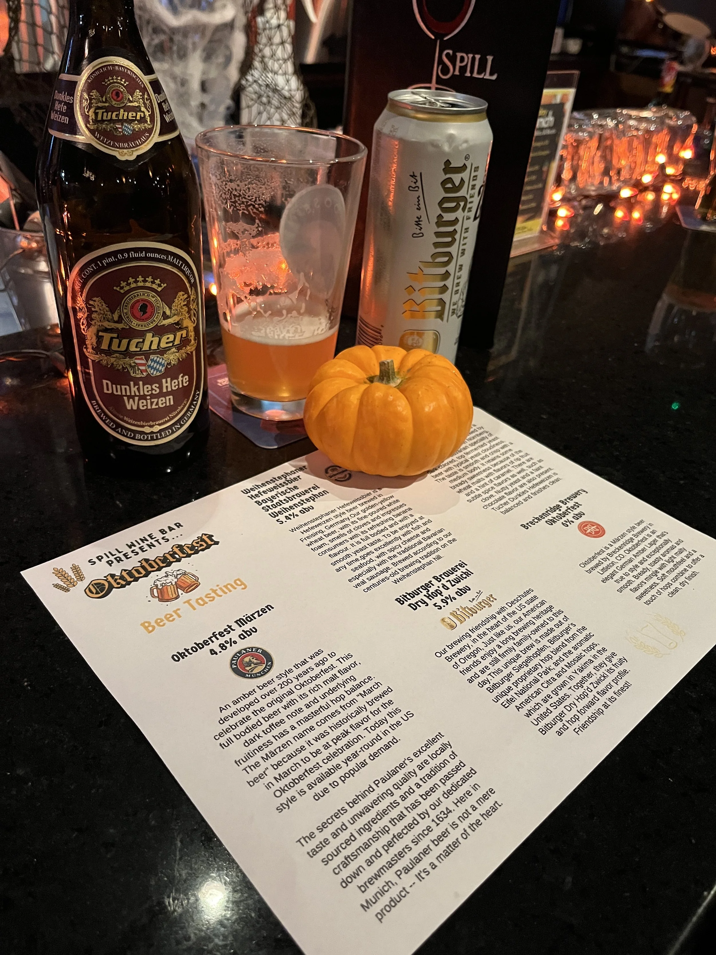 A beer tasting setup at a bar with a bottle of Tucher beer, a glass partially filled with beer, a can of Whitter Bier, a small pumpkin, and a printed beer tasting menu on a dark countertop.