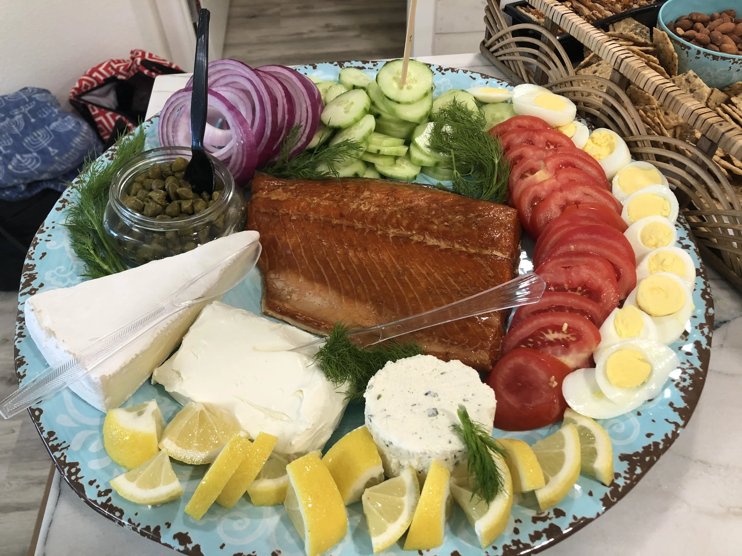 A plate with assorted foods including smoked salmon, sliced cucumbers, sliced tomatoes, boiled eggs, red onions, capers, lemon wedges, and various cheeses.