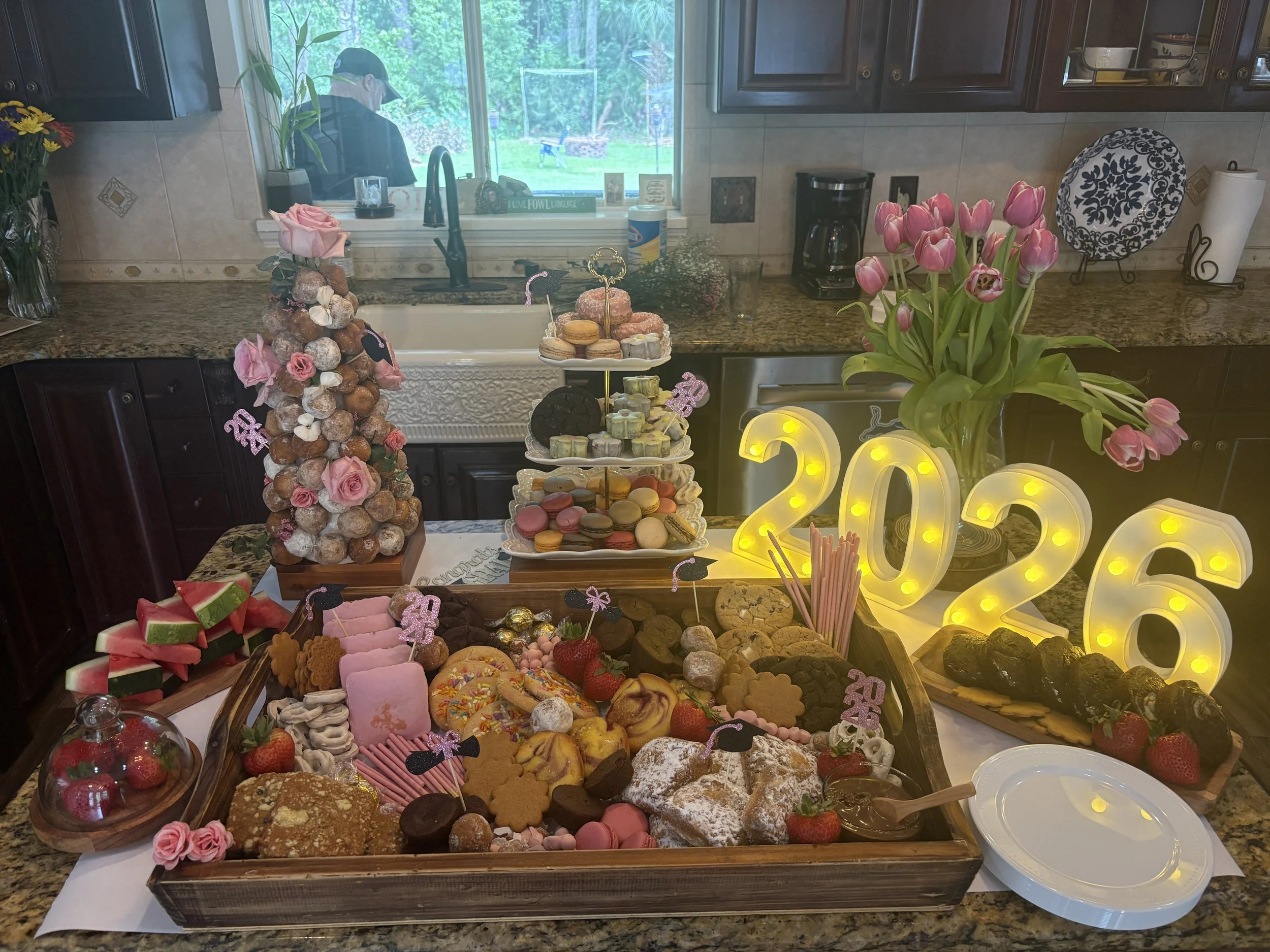 A kitchen with a table filled with various desserts, including cookies, cupcakes, macarons, strawberries, and watermelon slices. There are pink tulips in a vase and a lit decorative "2023" sign. A window in the background shows a person outside in a 
