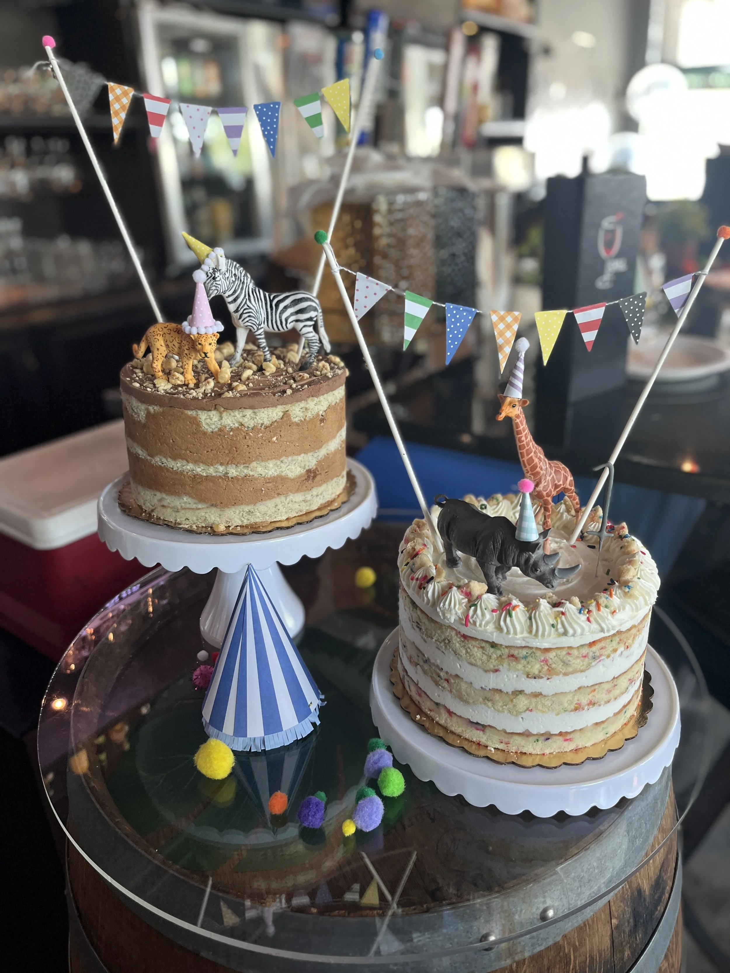 Two birthday cakes decorated with animal figurines, party hats, colorful bunting, and festive decorations on a glass platter.