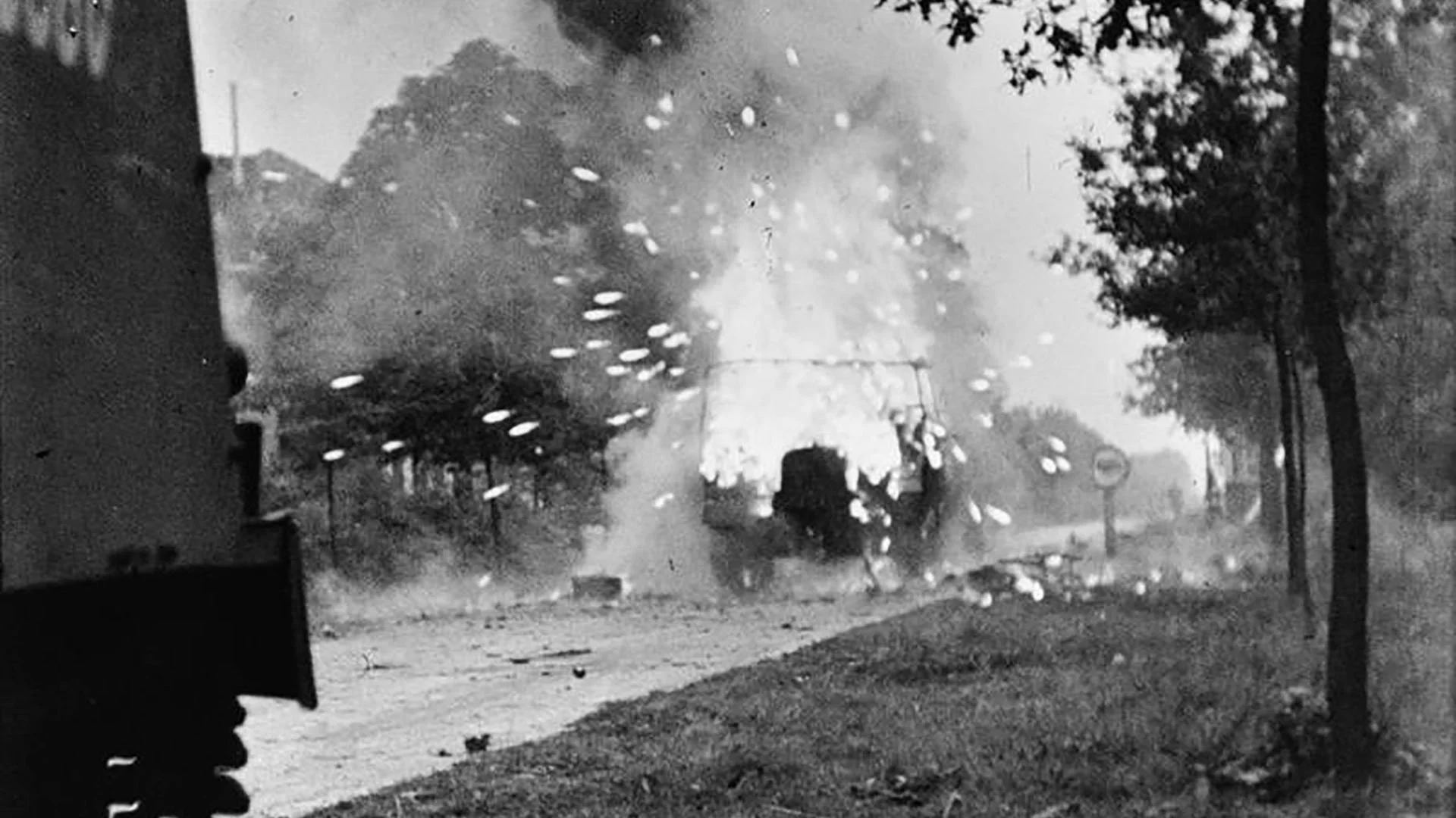 Black and white photo of a car exploding or crashing, with smoke and debris in the air, on a street lined with trees.