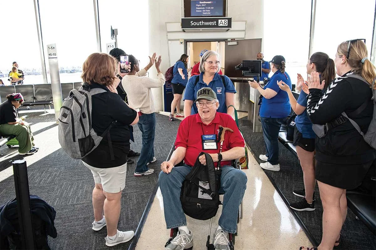 A group of people welcoming a smiling man in a wheelchair at an airport terminal gate labeled Southwest A8. The man is wearing a red polo shirt, a lapel badge, and a cap that says 'Army.' The scene appears celebratory, with several people clapping and taking photos.