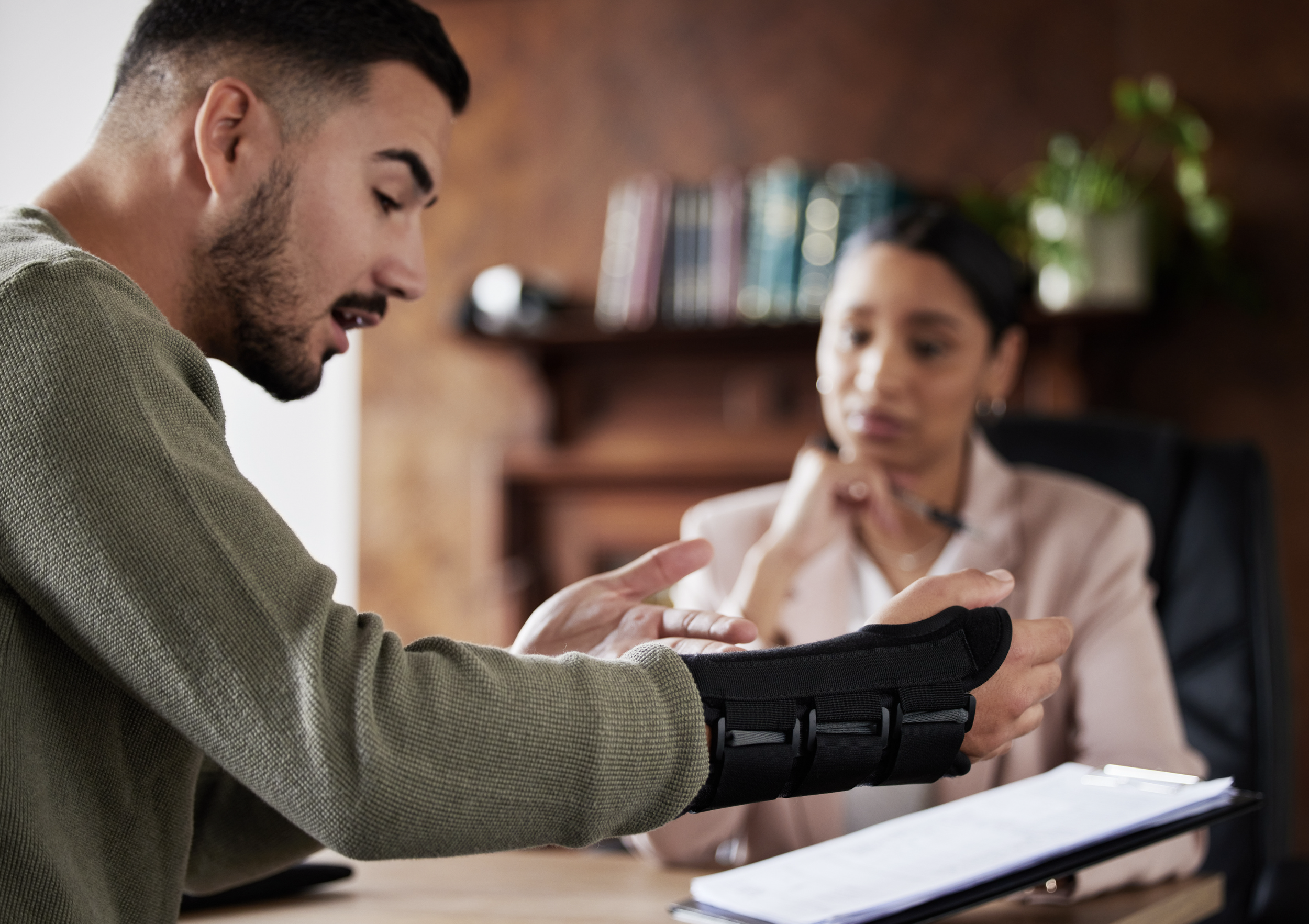 A young man with a cast on his arm showing it to a woman sitting at a desk in an office.