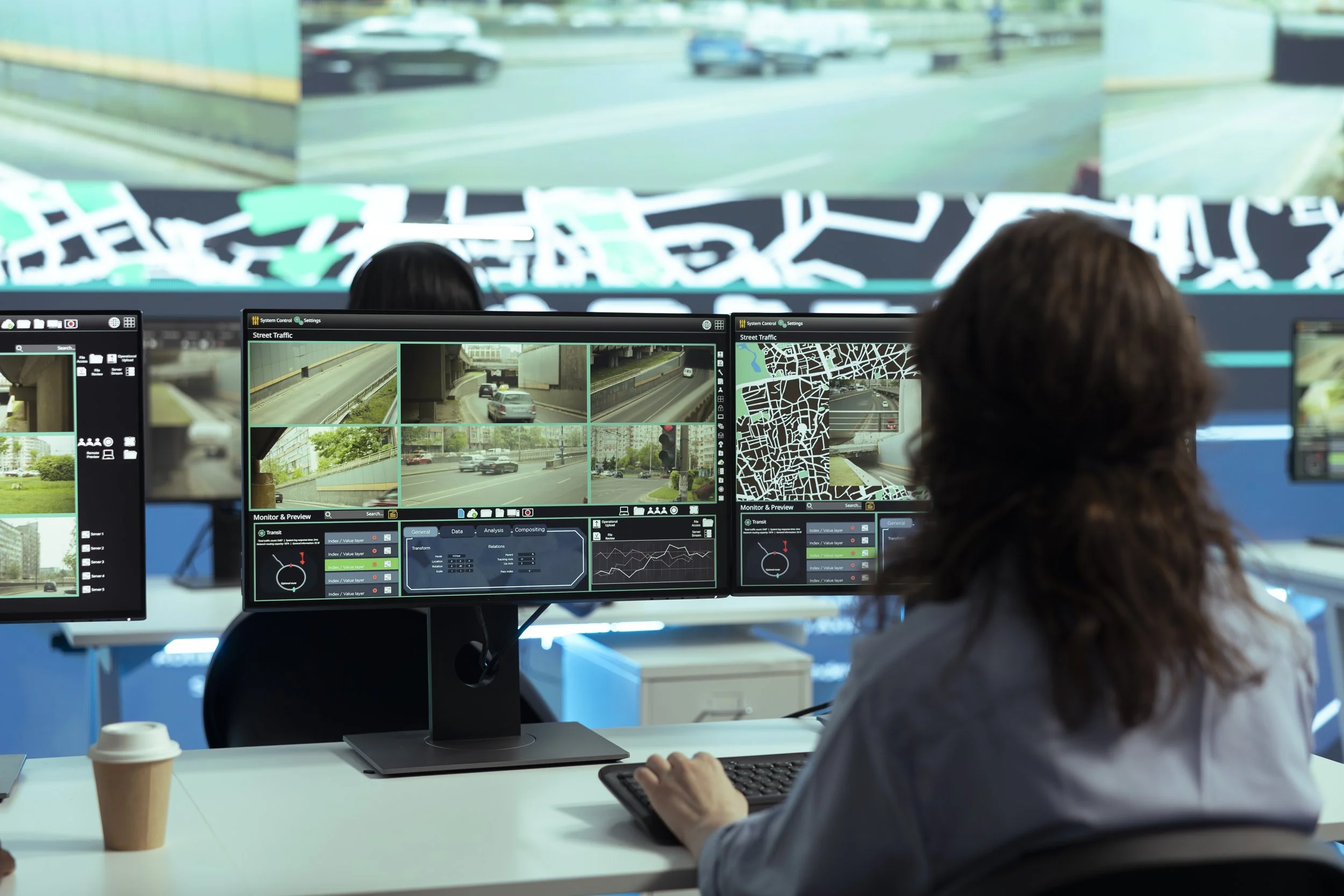 Control room with multiple monitors displaying live traffic footage and maps, woman with curly hair working at desk with coffee cup.