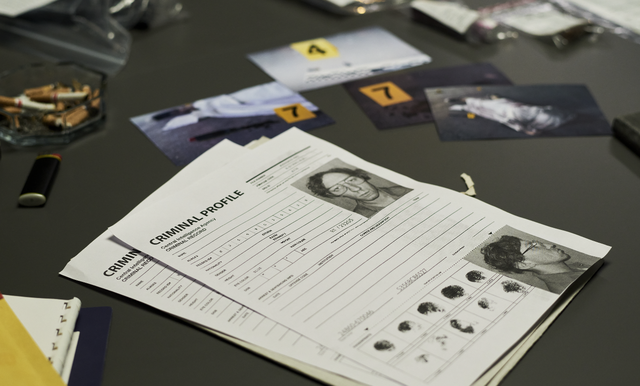 A police criminal profile report with a photo of a woman wearing glasses, placed on a desk with scattered photographs, bulletins, and miscellaneous items.