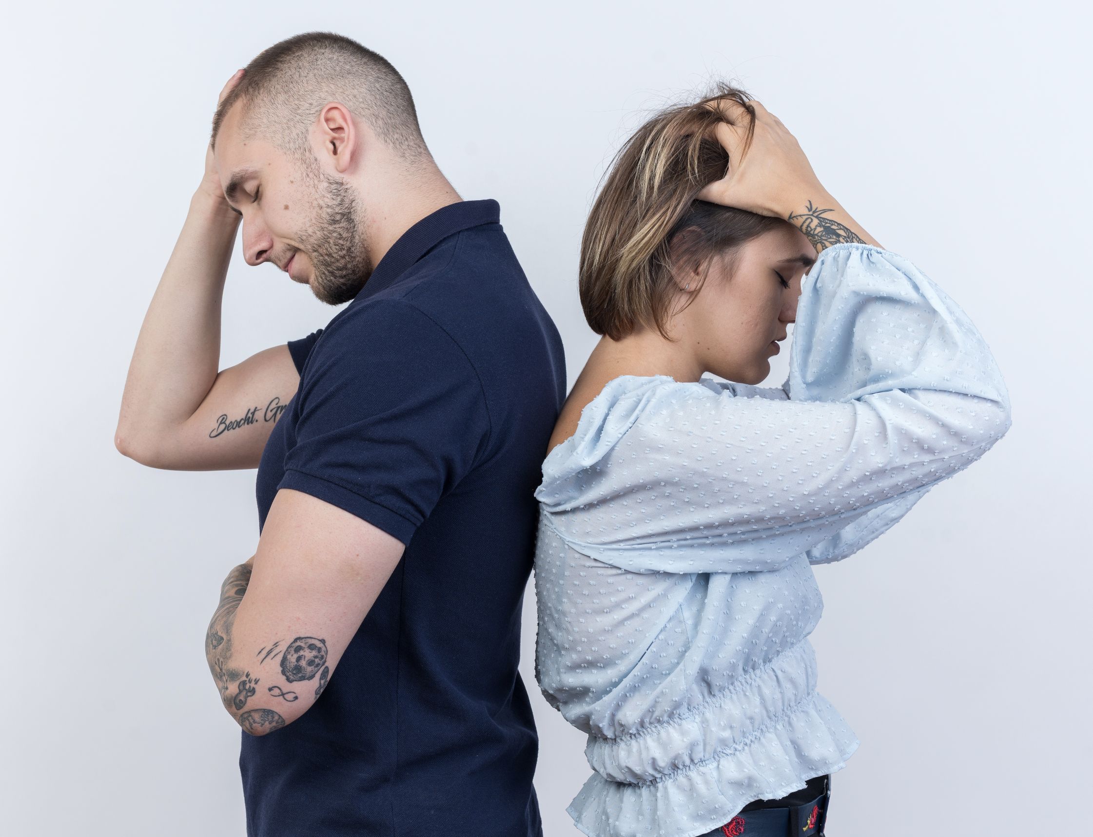 A man and woman stand back to back with their eyes closed, hands on their heads, appearing upset or stressed.