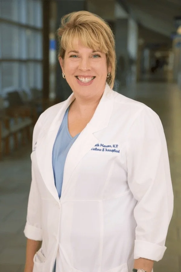 A smiling woman in a white medical coat standing in a hospital corridor.