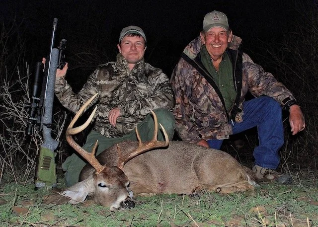 Two men in camouflage and outdoor clothing kneeling behind a large, dead buck with antlers, outdoors at night, one holding a rifle.