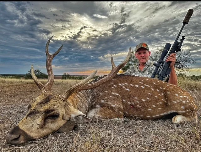 A hunter sitting behind a large deer with antlers, holding a rifle, in an open field under a cloudy sky at sunset.