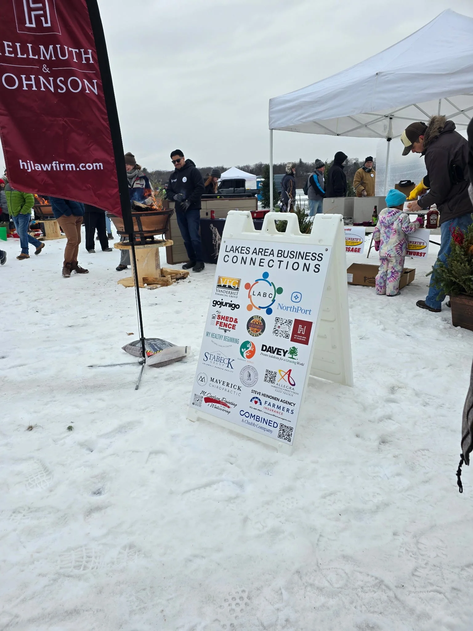 People attending an outdoor winter event, standing on snow, with booths and a signboard displaying various local business logos at Lakes Area Business Connections.