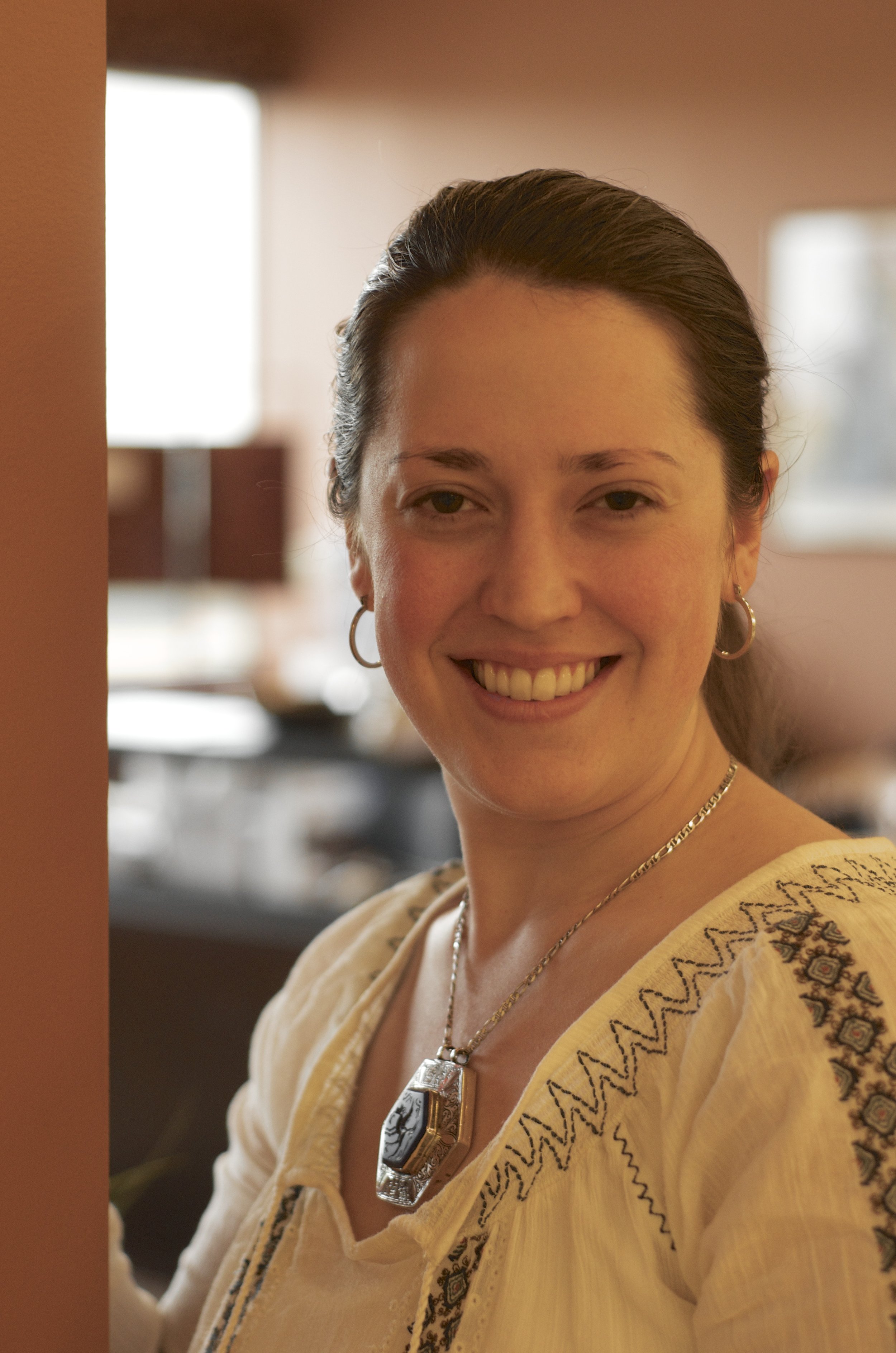 A woman smiling in an indoor setting, wearing earrings, a necklace with a large pendant, and a cream-colored embroidered blouse.