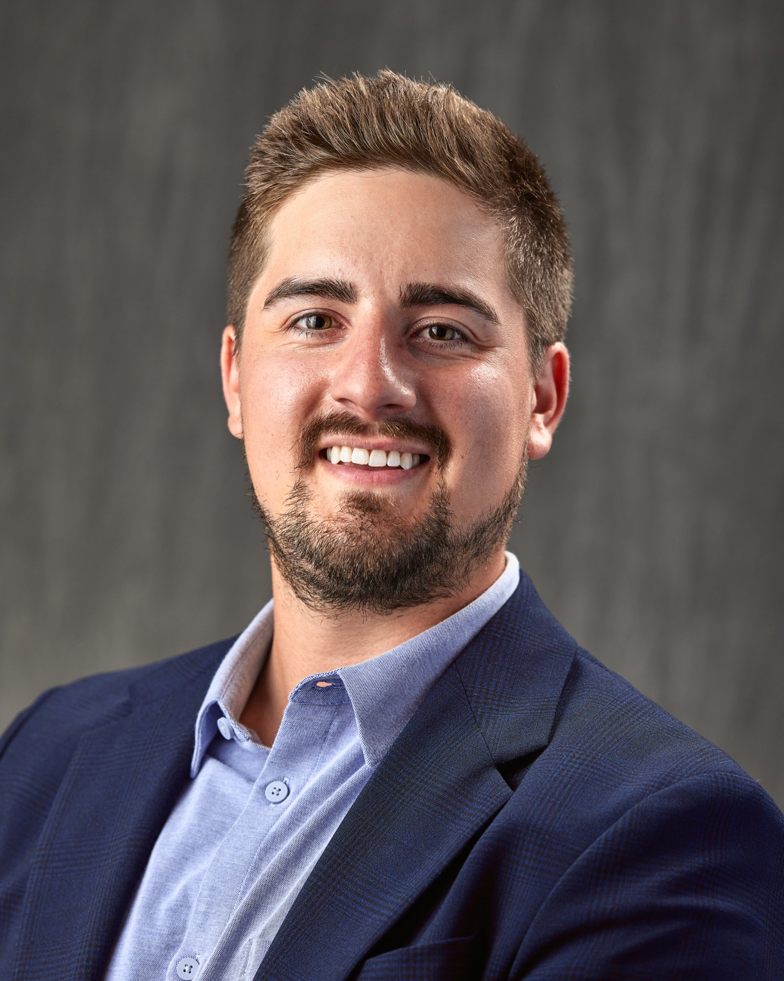 A headshot of a young man with short brown hair, a beard, and light skin, smiling, wearing a blue blazer and a light blue collared shirt, against a gray background.