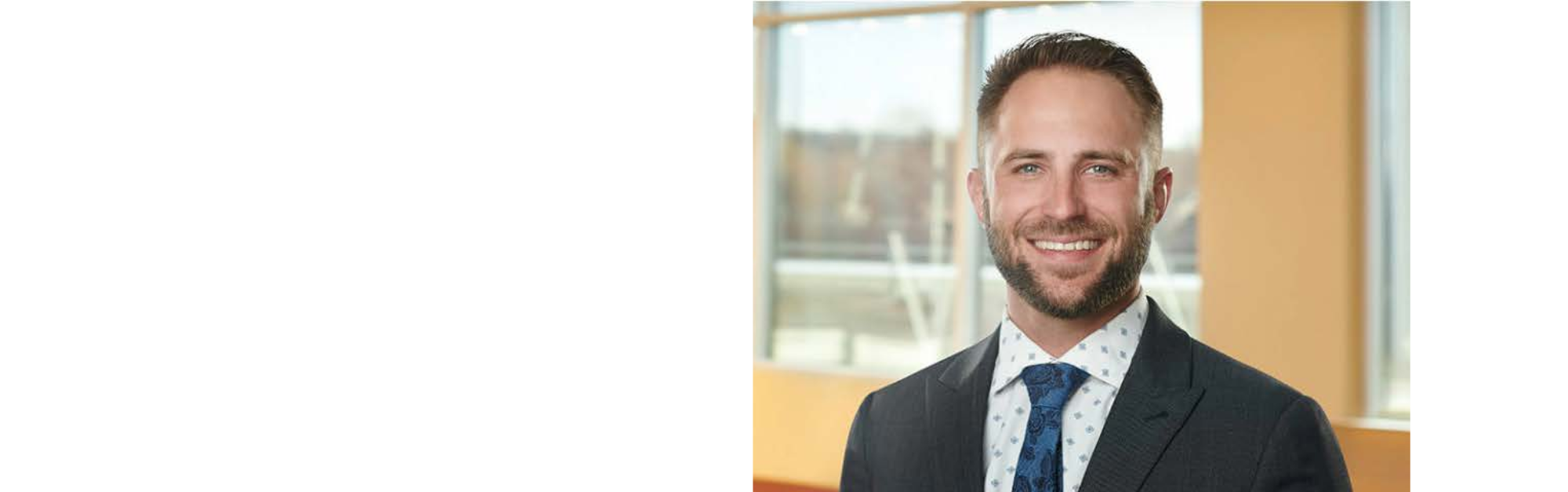 A smiling man with light brown hair and a beard, wearing a dark suit, white shirt with a pattern, and a blue tie, standing in front of large windows with a cityscape background.