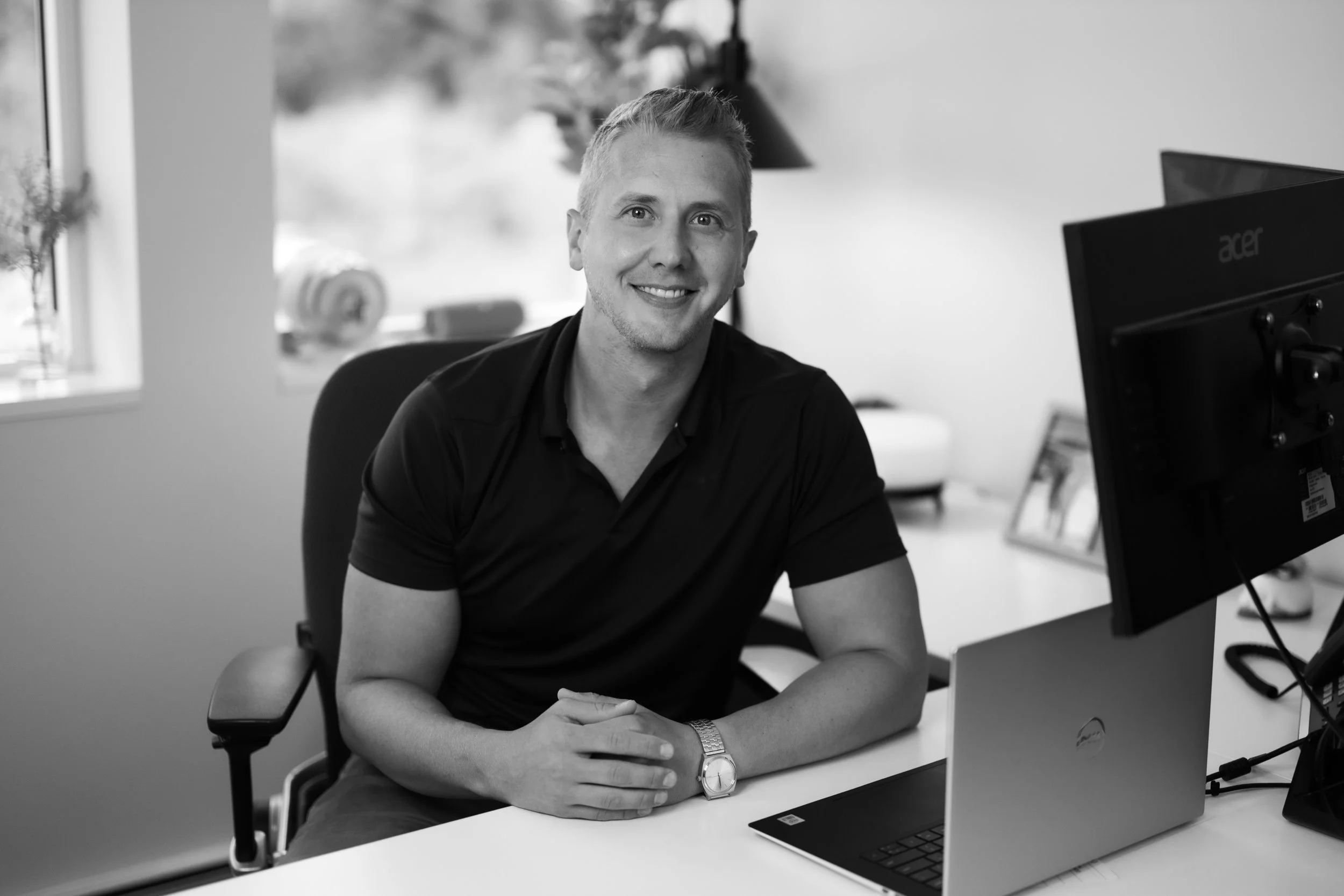 A smiling man sitting at a desk with a laptop and a computer monitor in a bright office.