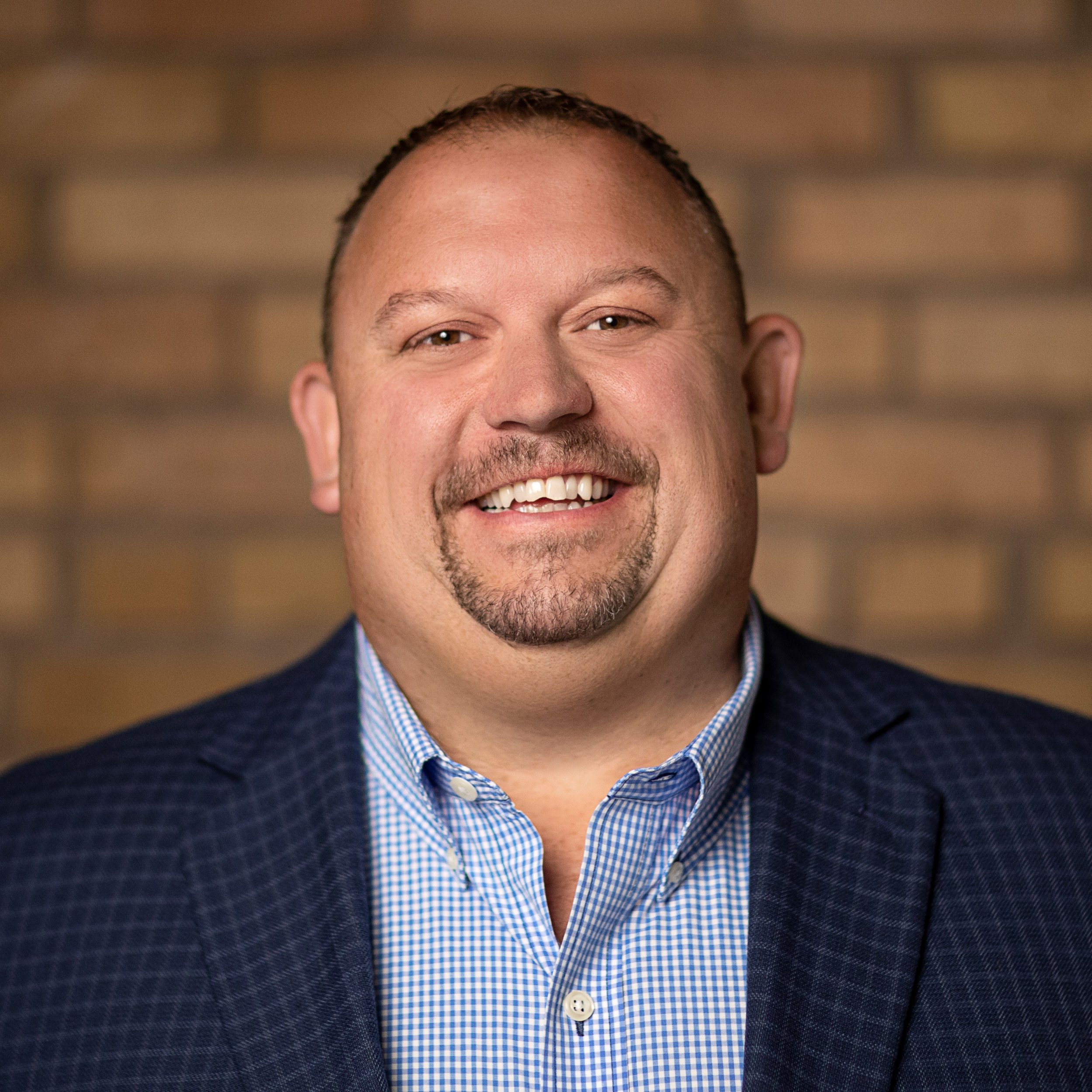 A headshot of a smiling man with short hair and a goatee, wearing a blue checkered shirt and a dark blazer, against a blurred brick wall background.
