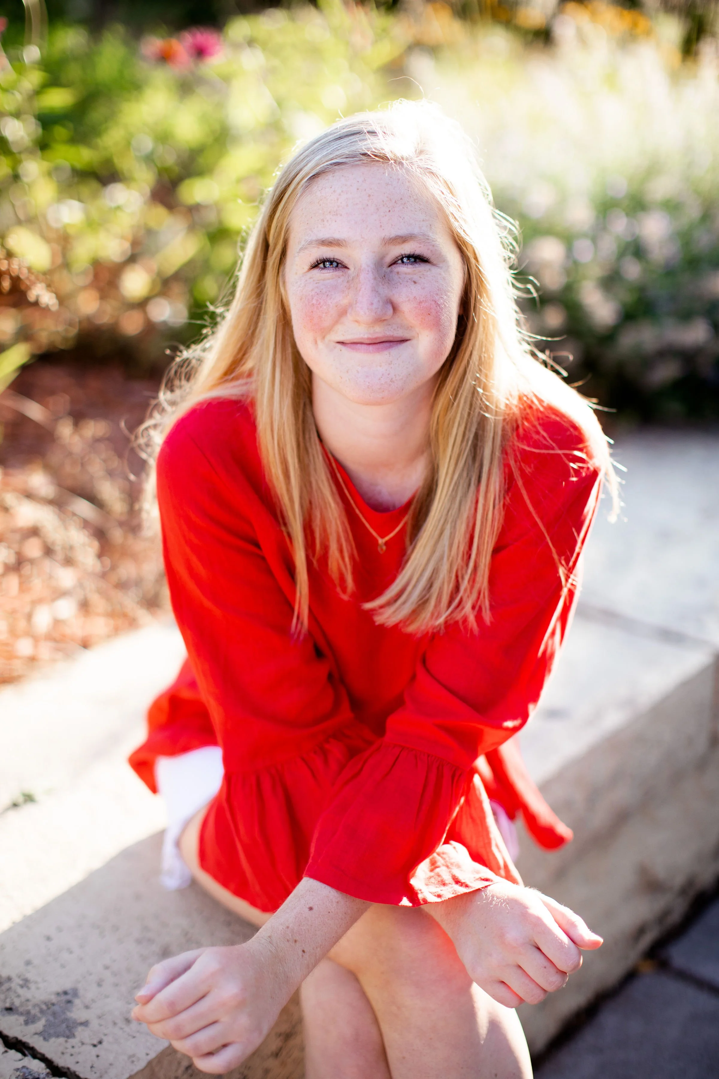 A young woman with red hair and freckles, smiling at the camera, wearing a red long-sleeve top, sitting outdoors on a stone ledge with greenery in the background.