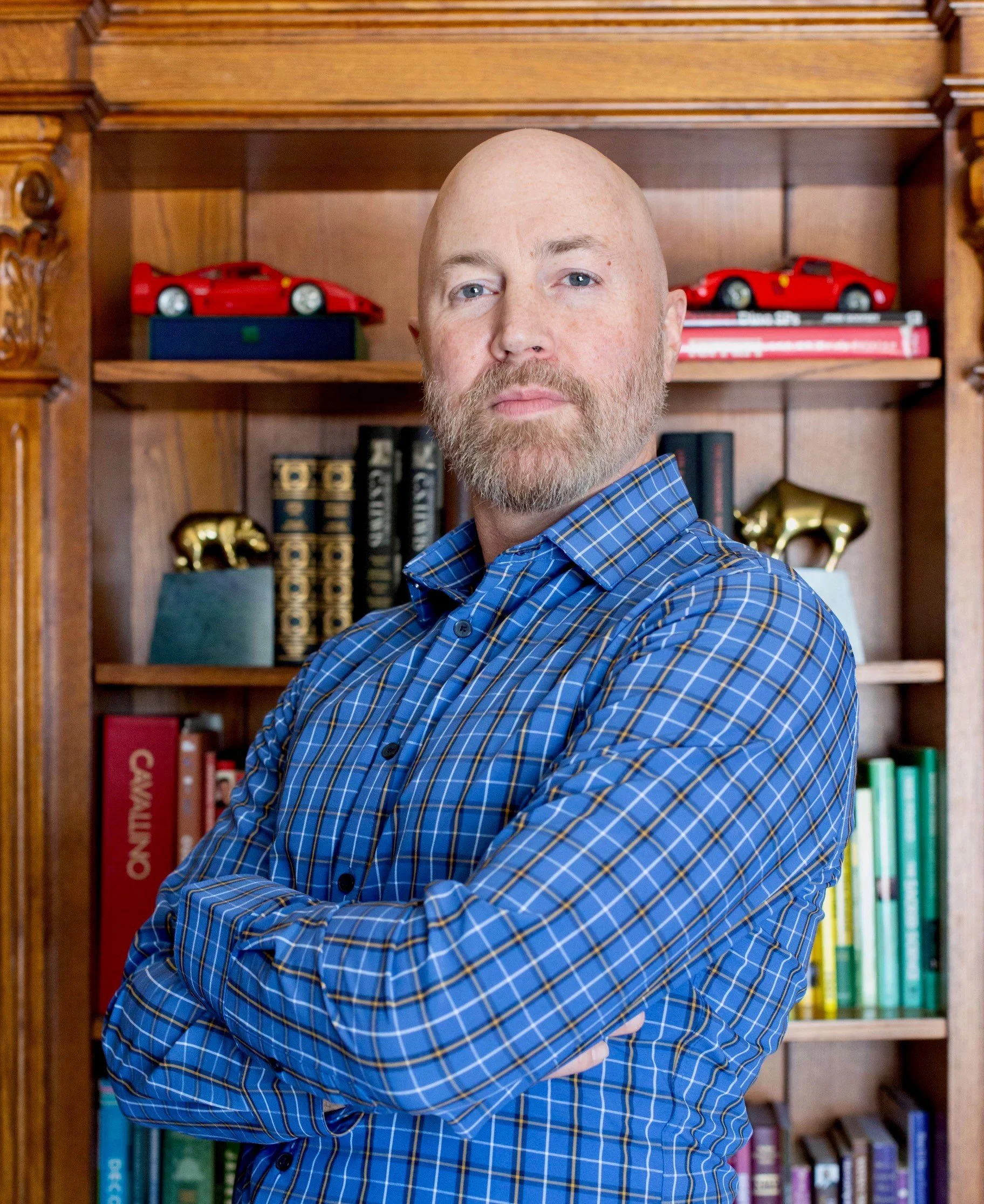 A man with a beard and bald head wearing a blue plaid shirt, standing with arms crossed in front of a wooden bookshelf decorated with model cars and books.