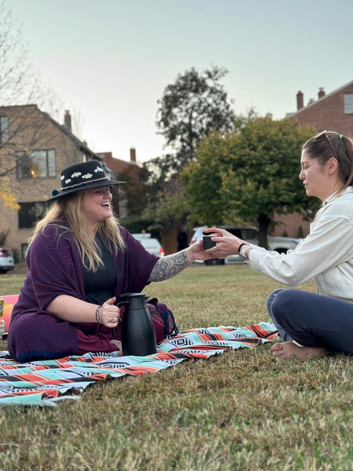 Two women sitting on a blanket outdoors, exchanging a small black container, with trees and houses in the background, during what appears to be a picnic or outdoor gathering.