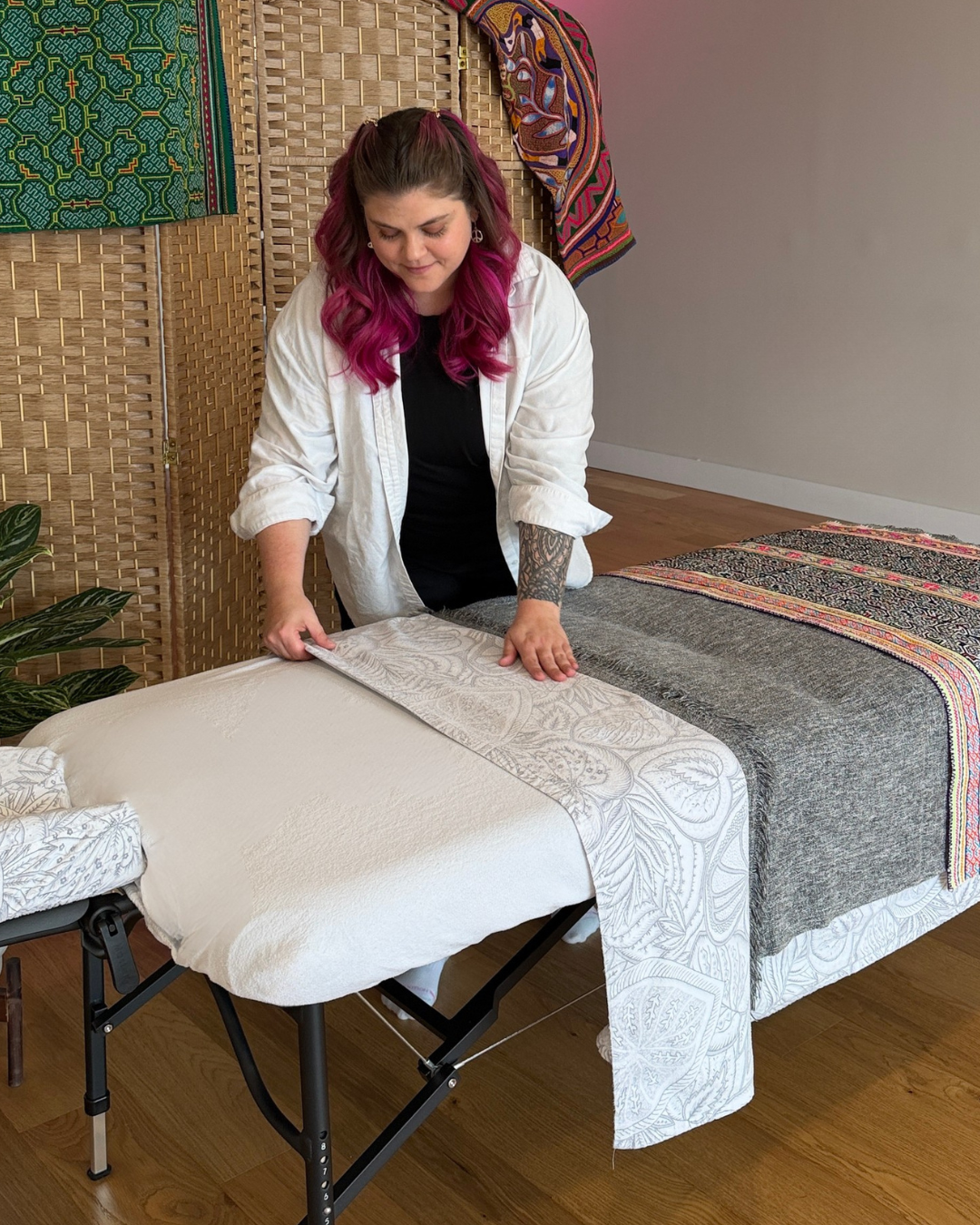 A woman with pink hair and a tattoo on her left arm is making a bed with white and patterned bedding in a room with wooden floors and a decorative folding screen. Colorful textiles hang on the screen.
