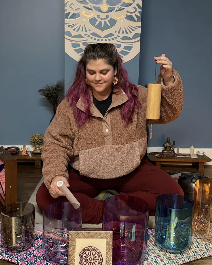 Woman with purple hair sitting cross-legged at a table with colored glass bowls, holding a wooden pendulum in one hand and a wand in the other, with spiritual objects and artwork in the background.