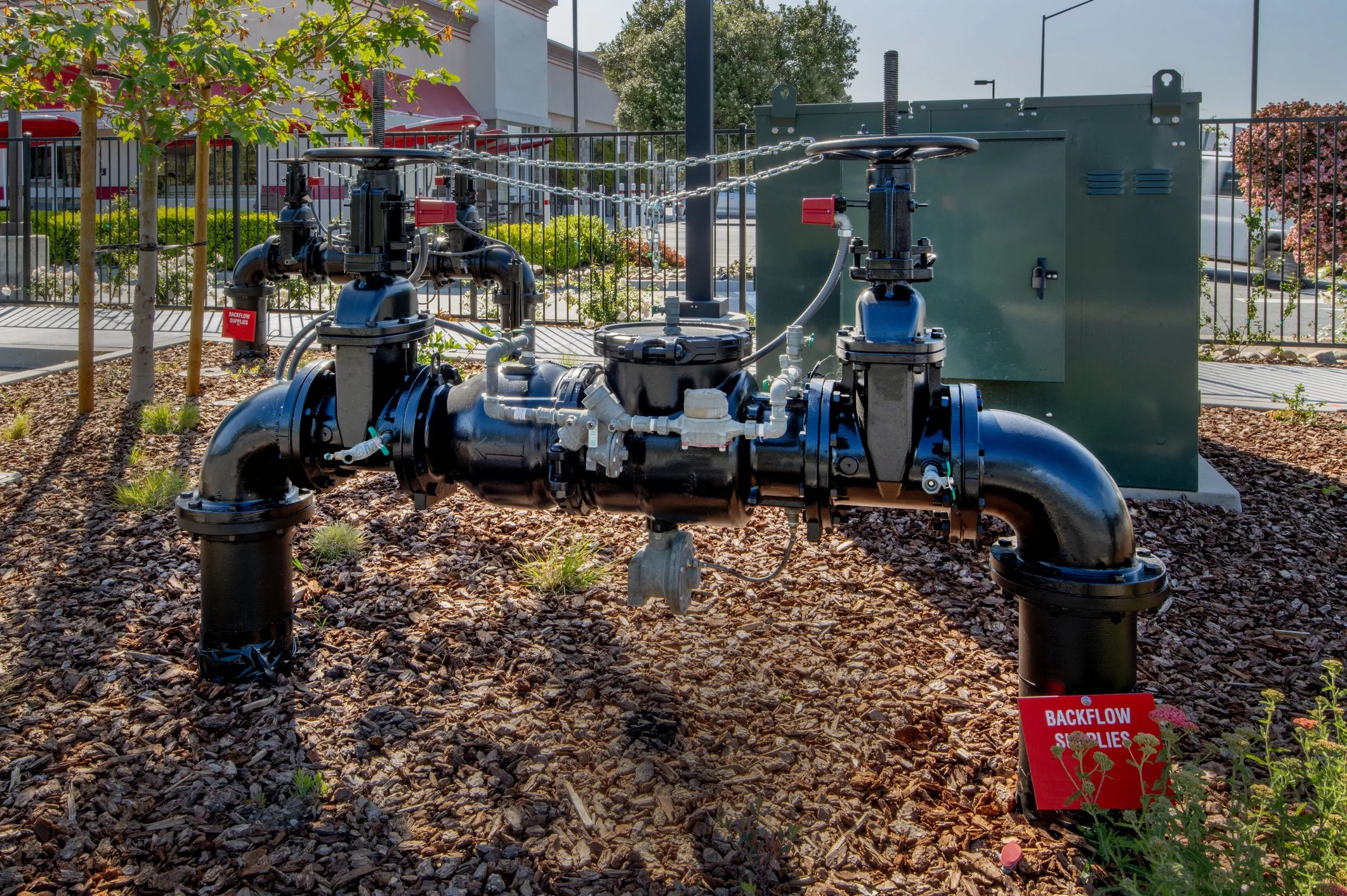 An outdoor view of a plumbing or irrigation system with black pipes, valves, and controllers installed on a landscaped area with mulch and small plants. There is a green electrical box or control panel in the background, surrounded by a black metal fence, and a sign with red text that says 'BACKFLOW SOLUTIONS.'
