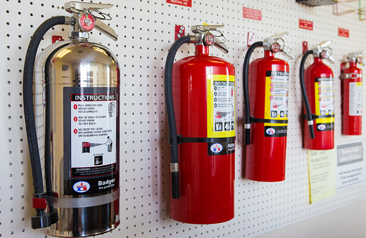 Multiple fire extinguishers mounted on a white pegboard wall, including a stainless steel and red models with instructions and labels.