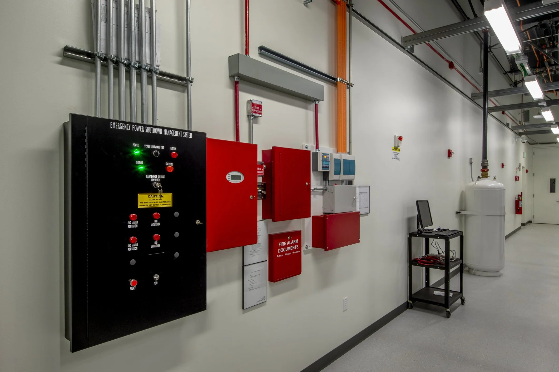 Industrial room with emergency power shutdown system, fire alarm documents box, and large gas tank along a white wall with ceiling-mounted lighting.