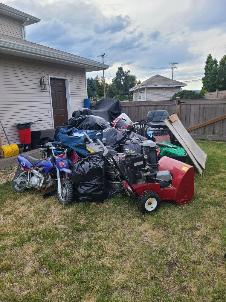 A pile of outdoor items including a snow blower, a small motorcycle, bags, and furniture on a backyard grass area near a house with a fence and another house in the background.