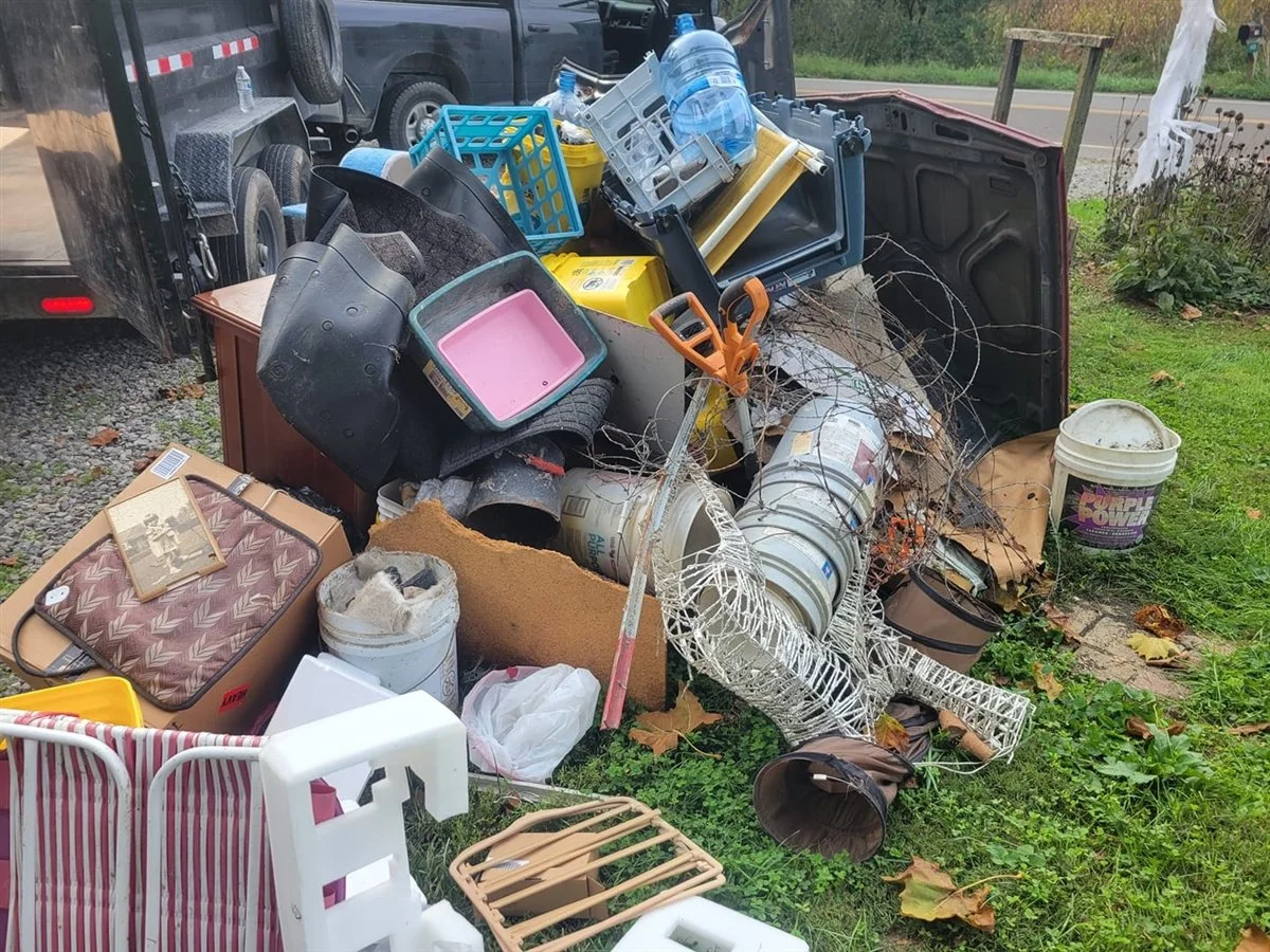 A truck bed overflowing with assorted trash and household items, including plastic containers, buckets, a ladder, a roll of wire, a chair, and other debris.