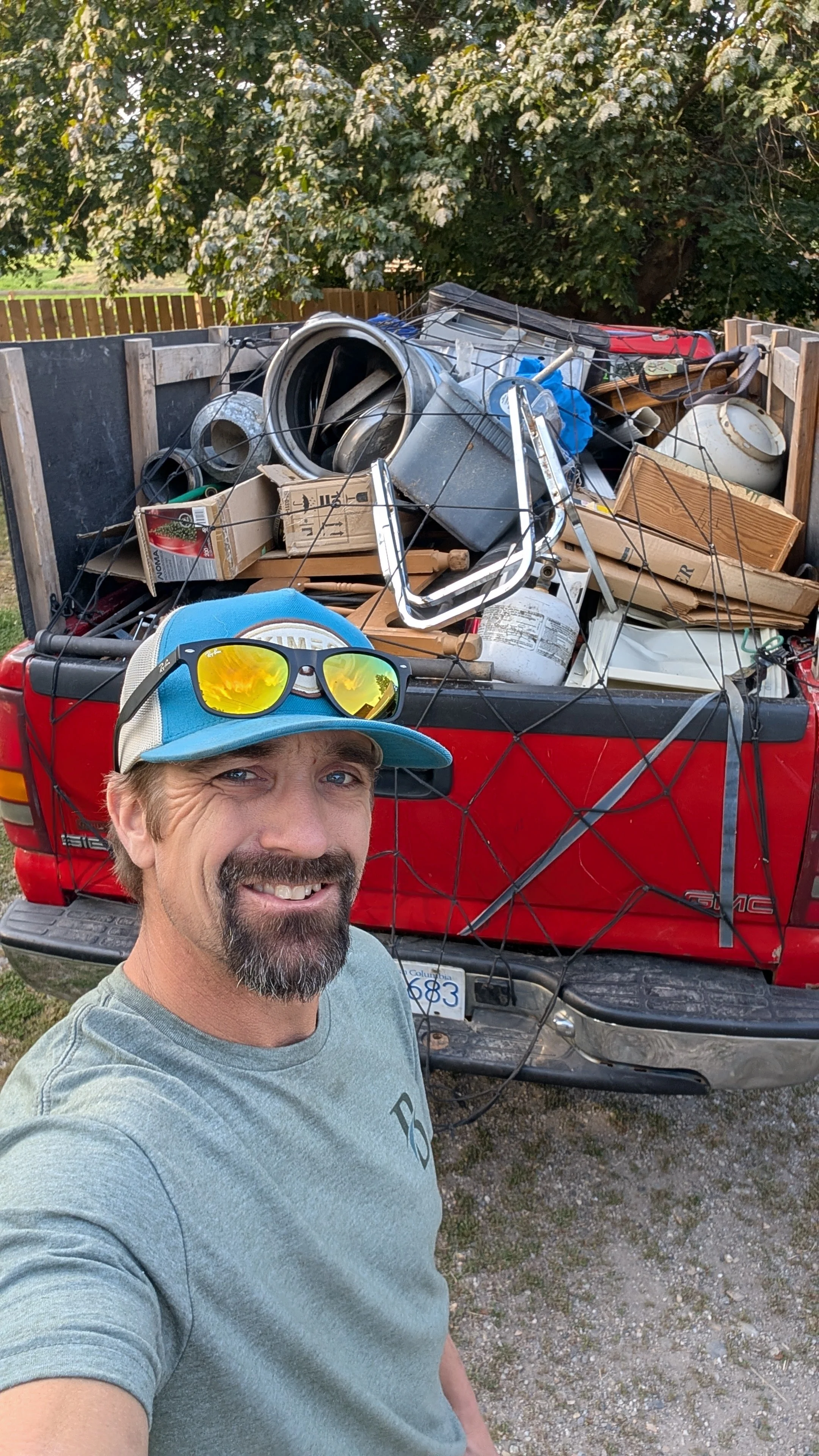 A man with sunglasses, a cap, and a goatee smiling for a selfie in front of a red pickup truck loaded with scrap metal and cardboard.
