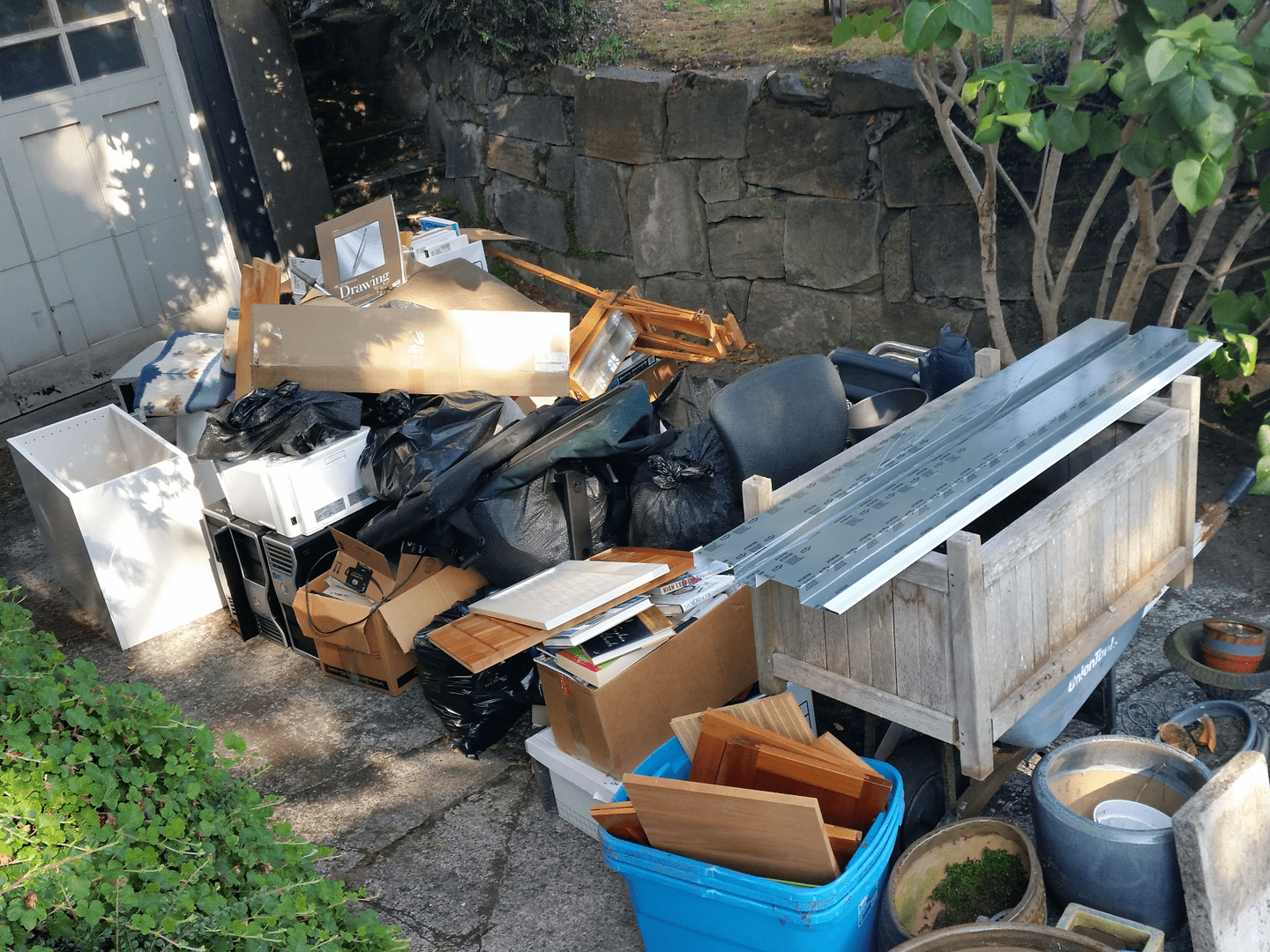A pile of assorted household items including furniture, electronics, boxes, plastic bags, and gardening materials outdoors on concrete pavement next to a stone wall and bushes.