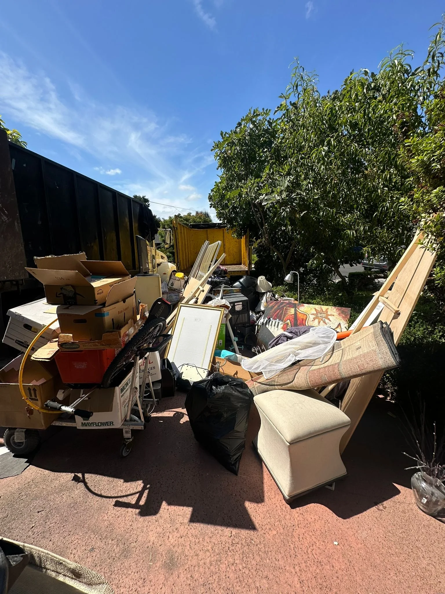 A cluttered outdoor space with various household items, furniture, and boxes piled on a concrete patio under a blue sky with some clouds.