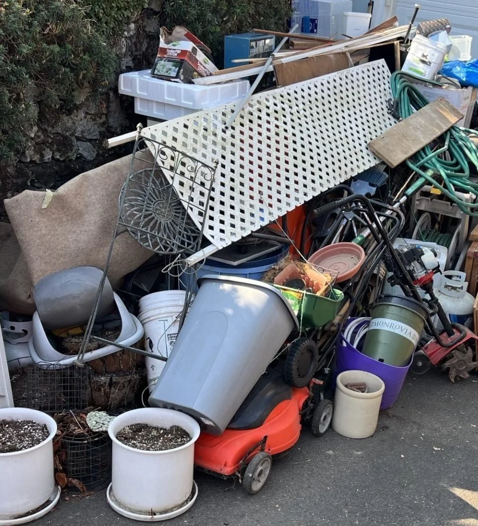 A large pile of assorted yard and garden tools, containers, and miscellaneous items haphazardly stacked outdoors on pavement, including plant pots, a lawnmower, hoses, buckets, and furniture.