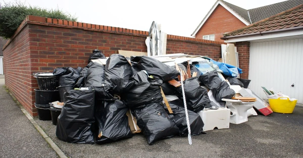 A large pile of trash and discarded household items, including black garbage bags, buckets, toilet bowls, and miscellaneous debris, stacked against a brick wall in a driveway.