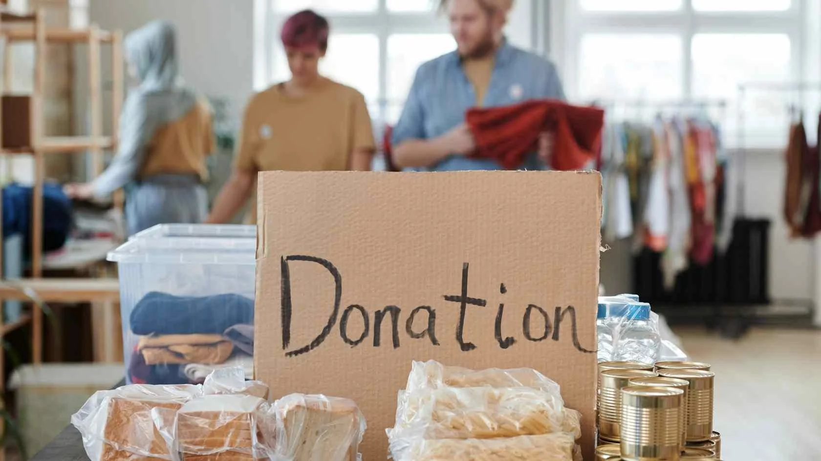 Table with donation items including canned goods and packaged food, with a handwritten 'Donation' sign, and three people in the background organizing clothing in a room.