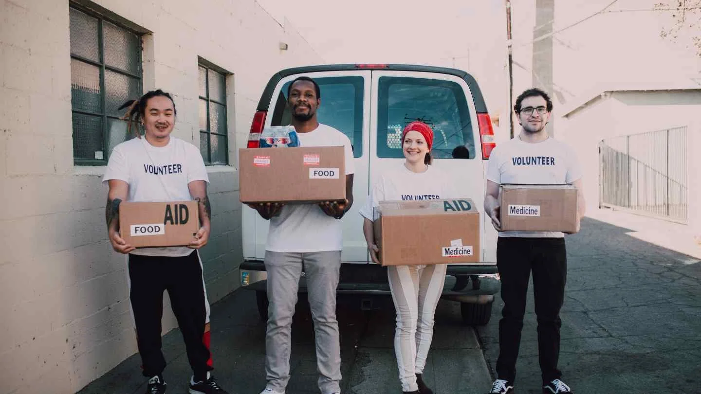 Four volunteers standing in front of a van holding boxes labeled with aid, food, medicine, and food, standing outdoors near a beige brick wall.