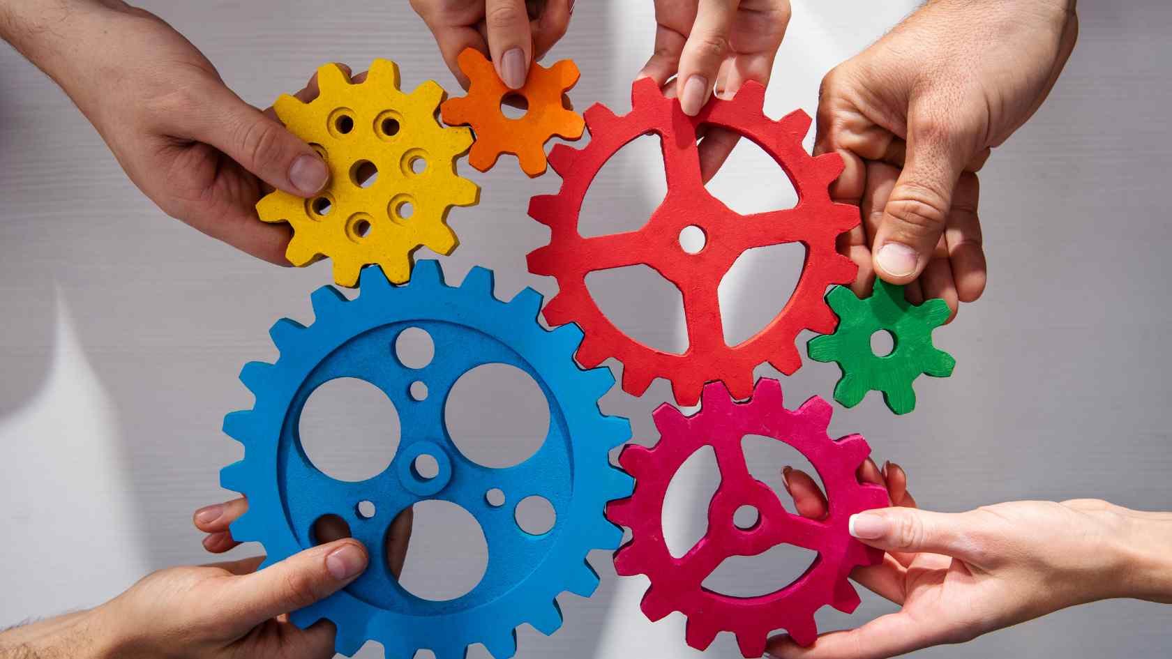 Multiple hands holding colorful interlocking gears made of plastic in yellow, orange, red, green, blue, and pink, suggesting teamwork or collaboration.