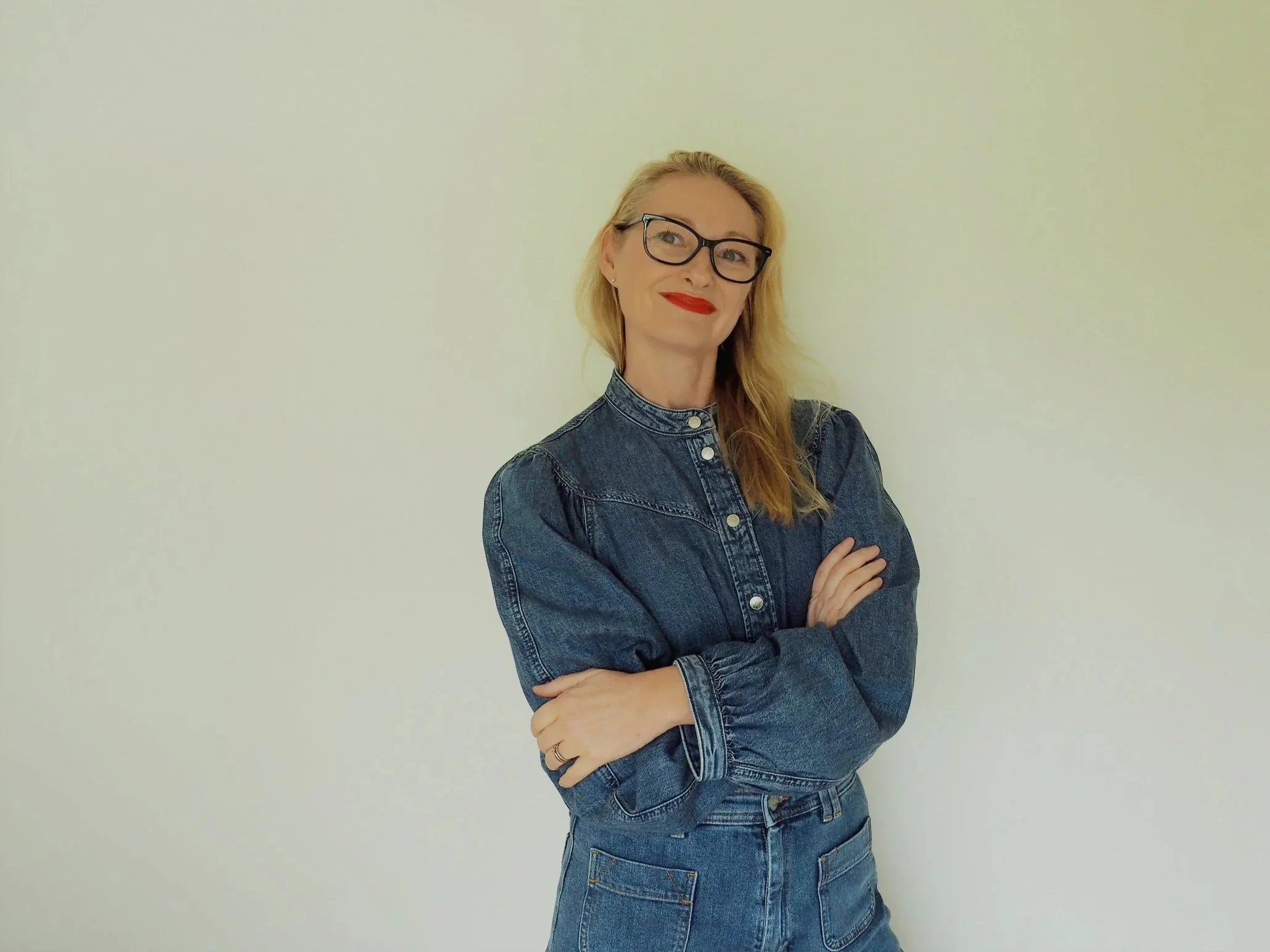 Woman with blonde hair, glasses, and red lipstick stands against a plain off-white wall. She is wearing a denim jacket and jeans, with her arms crossed. She is a Online Business Manager (OBM) and Virtual Assistant (VA)