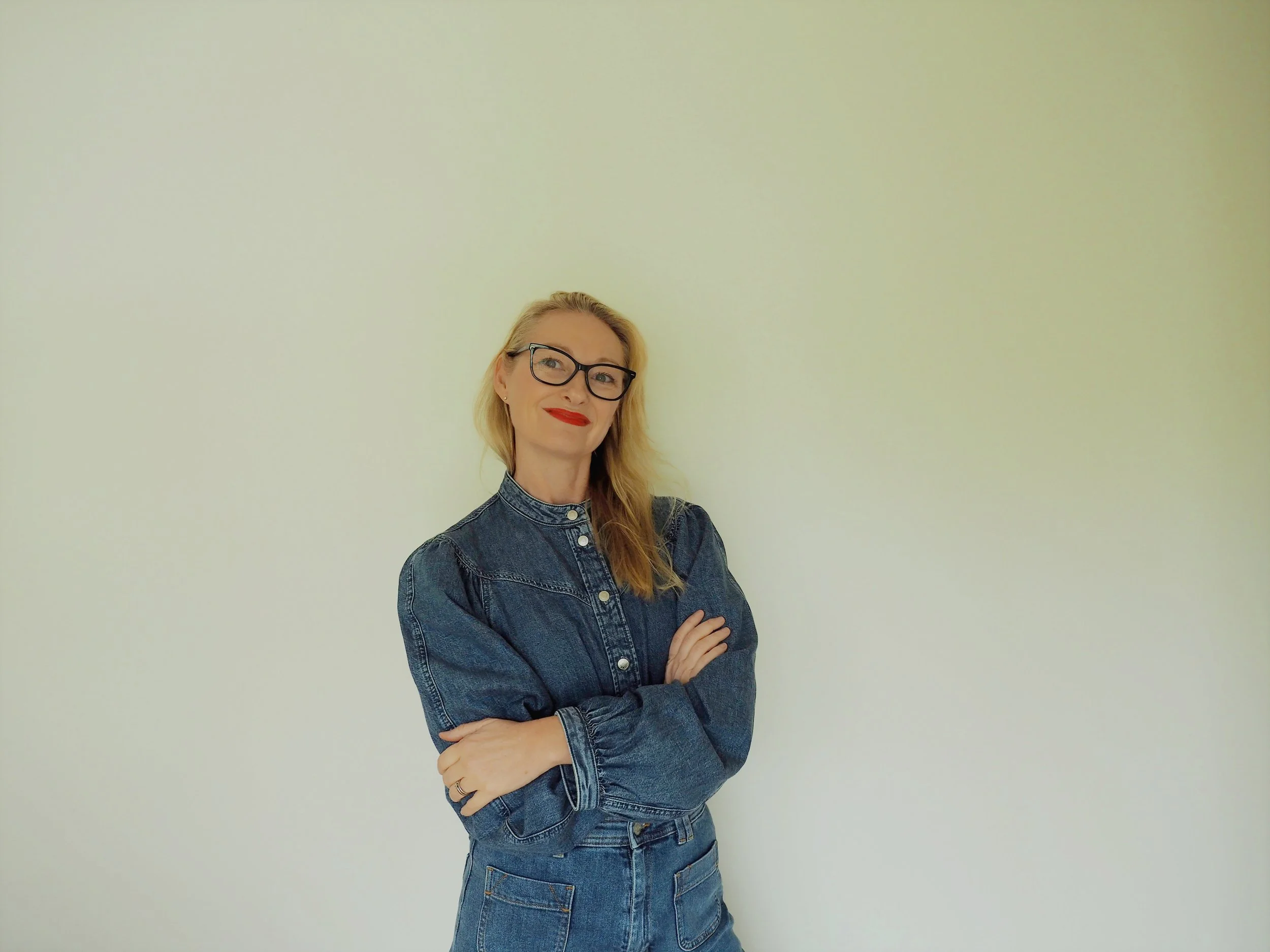 A woman with blonde hair, black glasses, red lipstick, wearing a denim jacket and jeans, standing with arms crossed against a plain light-colored wall.