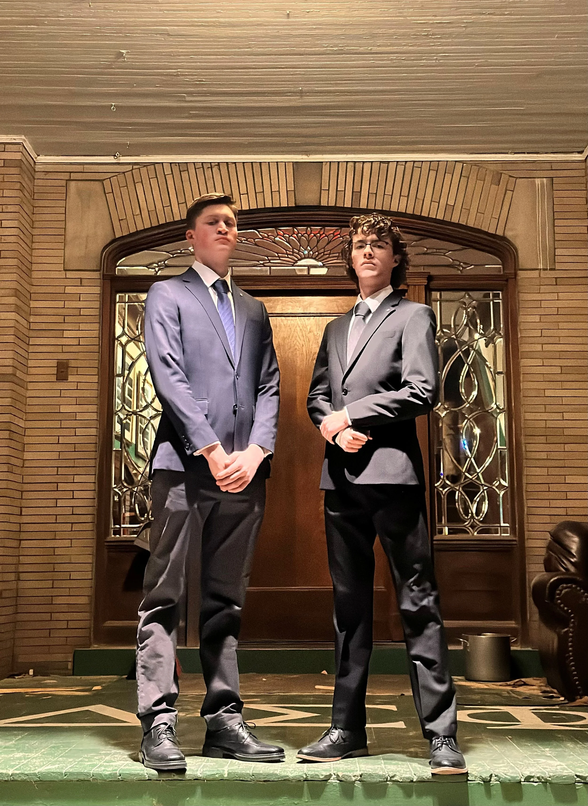 Two young men in gray suits stand confidently in front of a wooden door with stained glass windows in a brick-walled room.